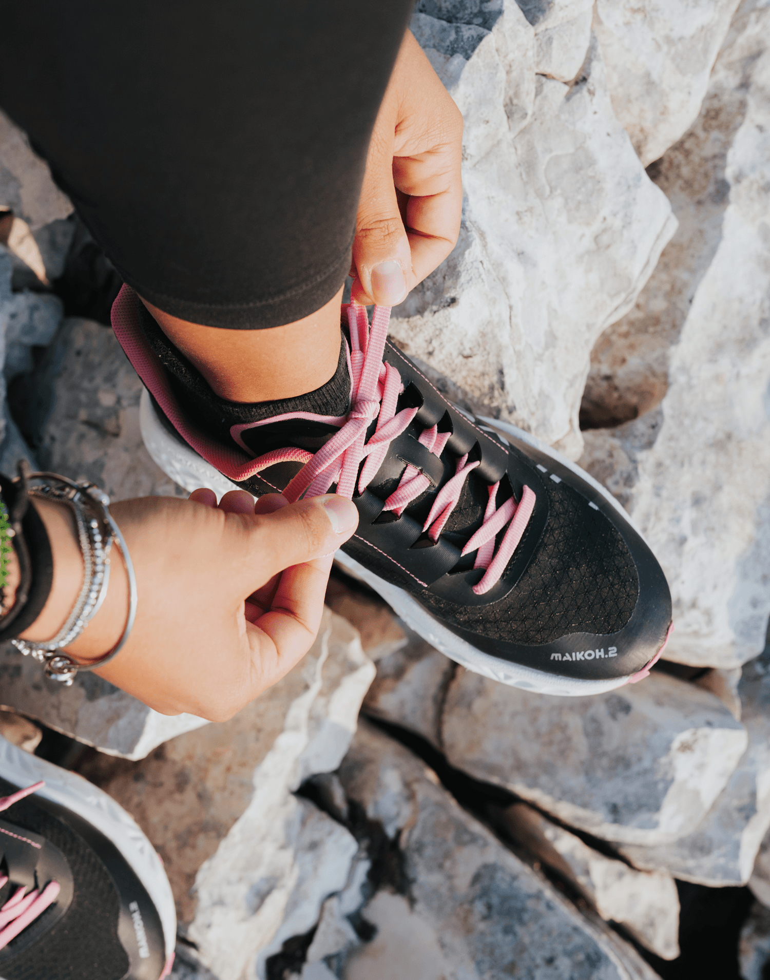 Overhead view of hands tying the pink laces of a black Maikoh.2 trail shoe on rocky terrain, with a second pair visible in the foreground.