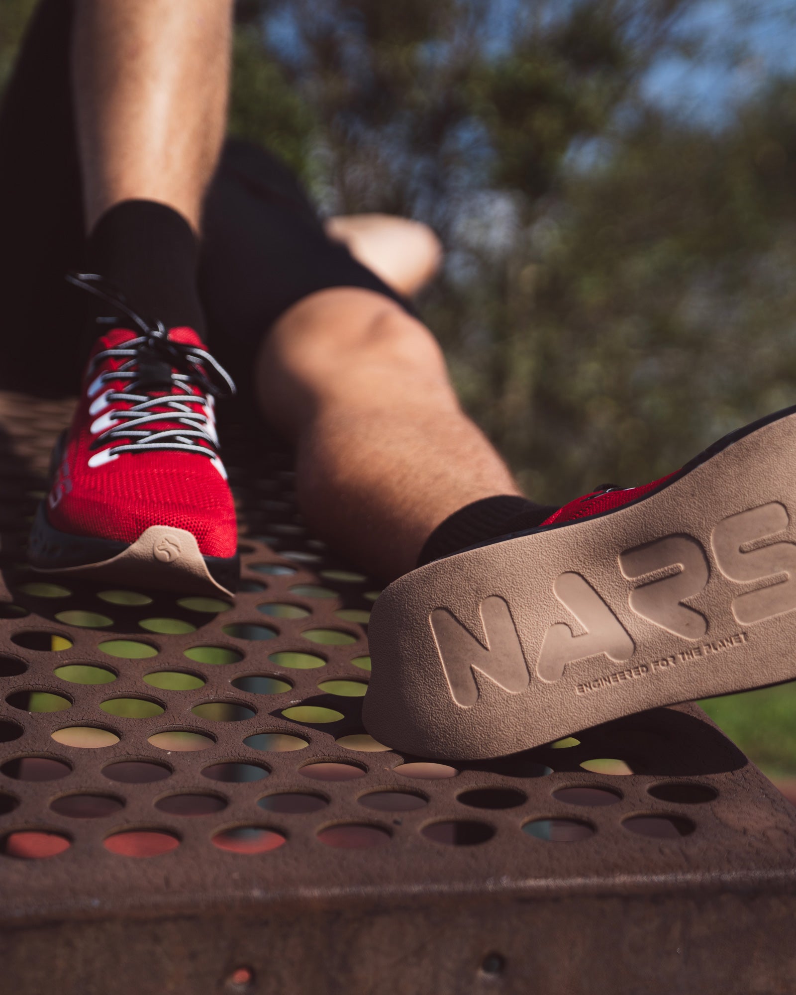 Close-up of a runner resting on a perforated metal surface, wearing red Ulysses running shoes with black laces and beige soles embossed with the word “NARS – Engineered for the Planet.”