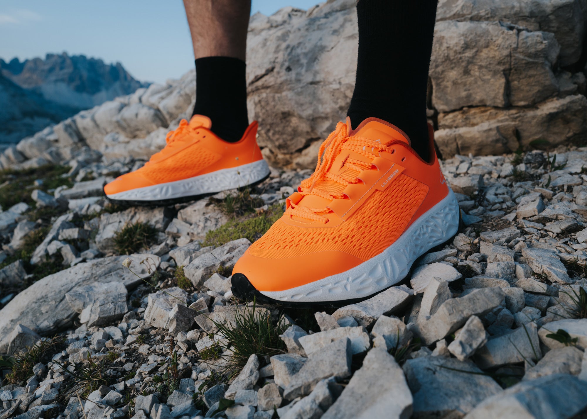Close-up of bright orange Ulysses trail shoes on a person's feet, standing on rocky terrain with mountains in the background.
