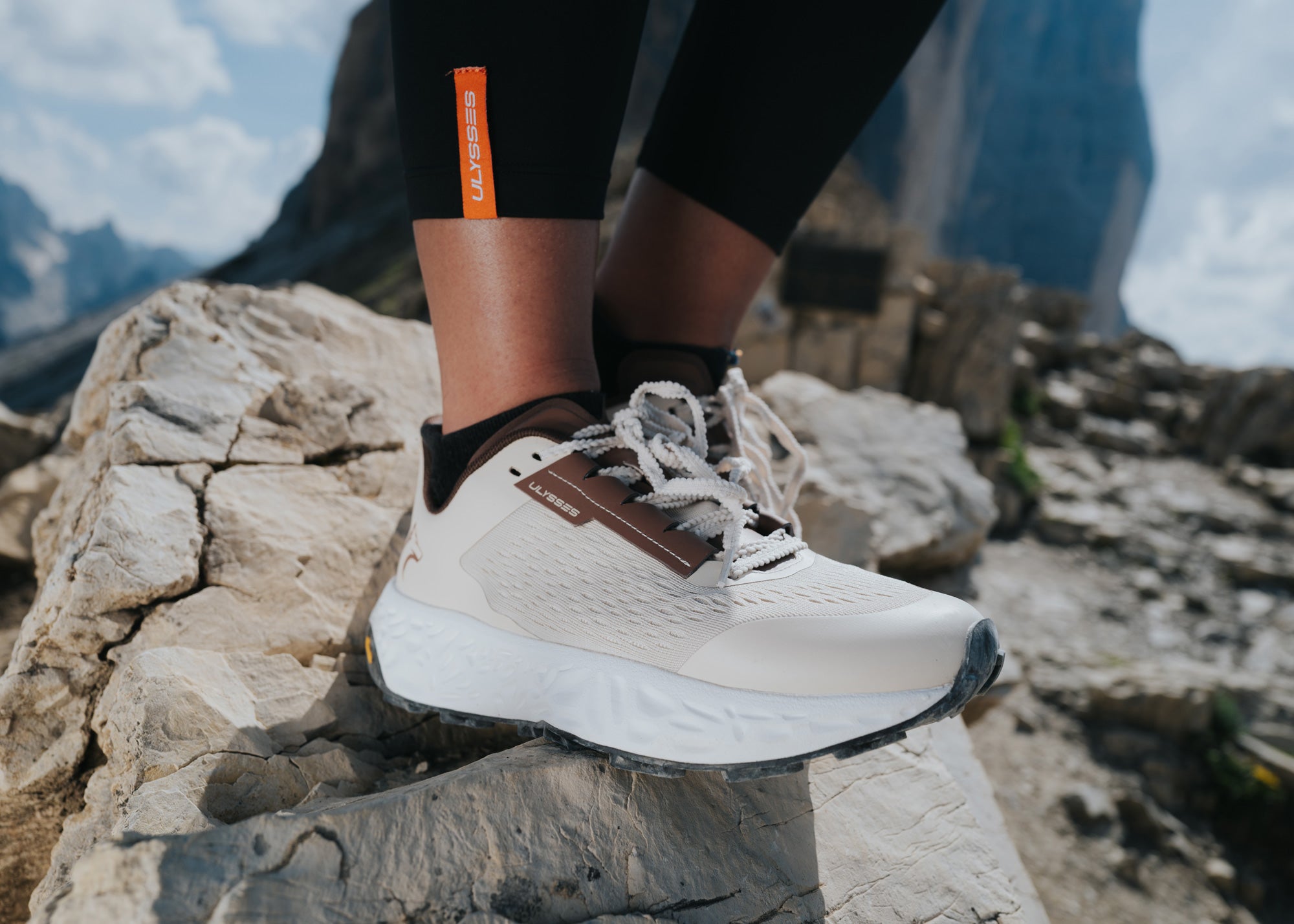 Close-up of a beige and brown Ulysses trail shoe on a woman's foot, standing on rocks with mountain scenery and an orange "ULYSSES" tab on her leggings.