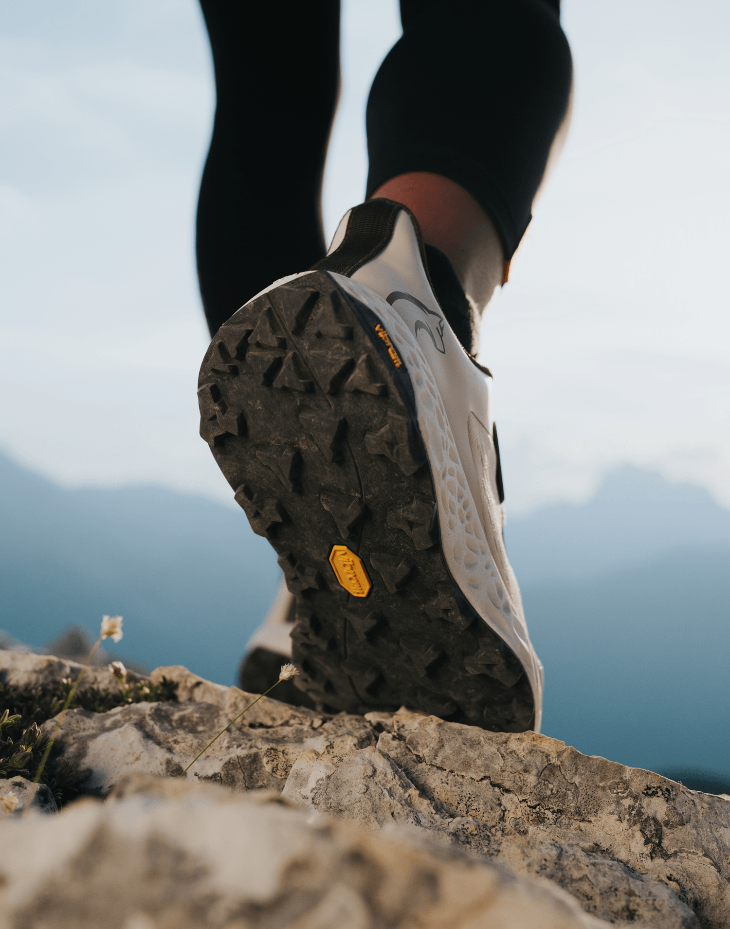 Close-up rear view of a Vibram-soled trail shoe mid-stride, showing the aggressive lug pattern against a mountain backdrop.