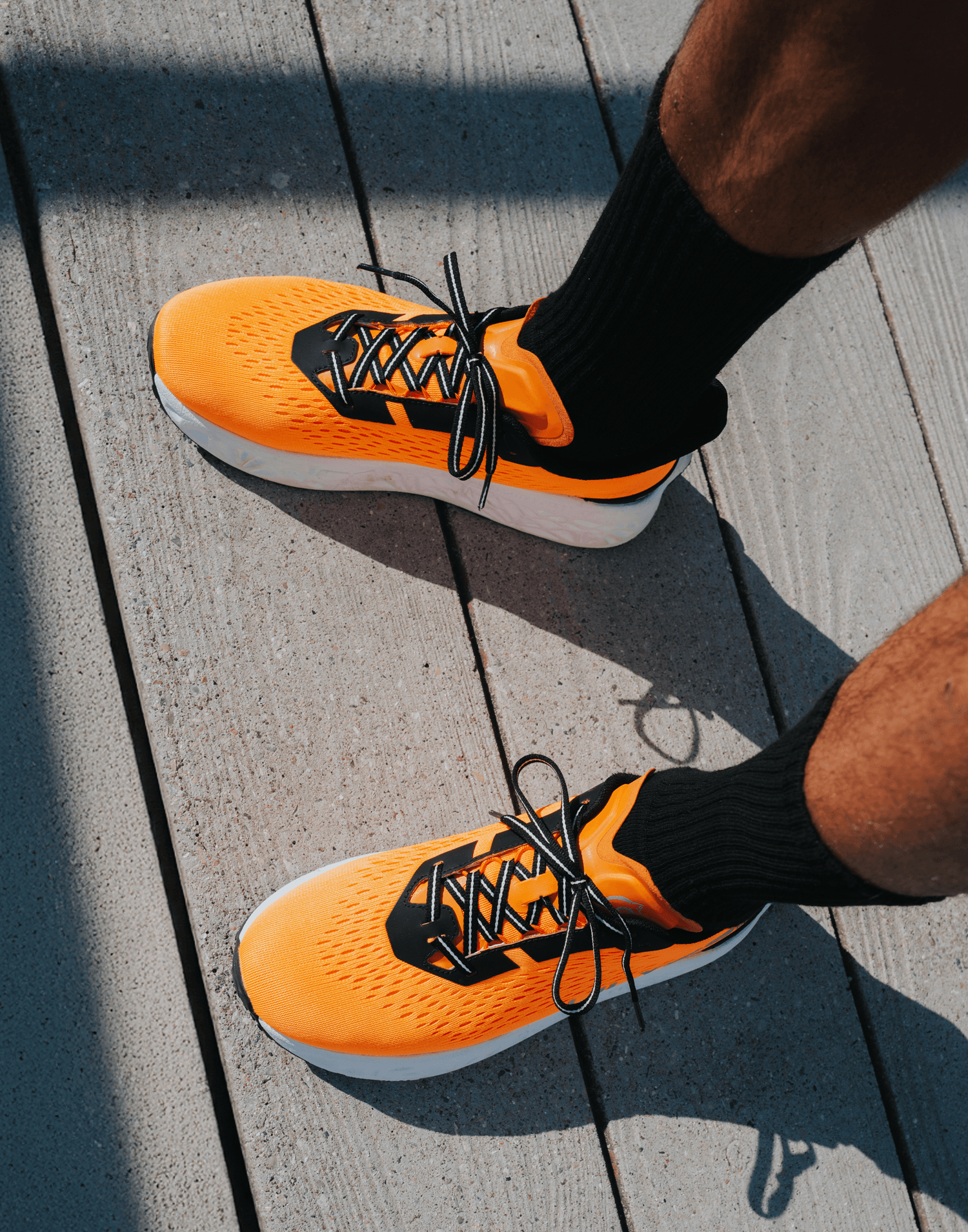 Overhead view of a runner's feet in orange Ulysses shoes with black laces, standing on a wooden deck in the sun.