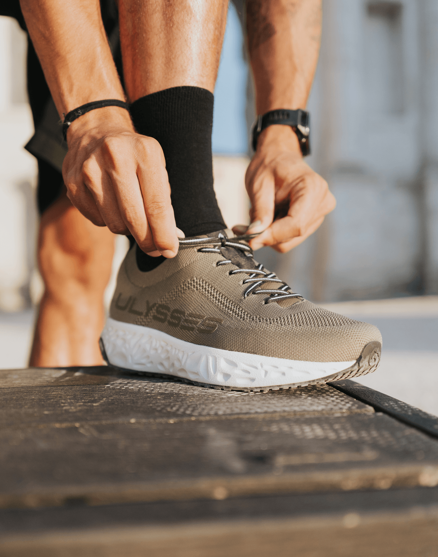 Close-up of hands tying the laces of an olive green Ulysses knit shoe, with a watch and bracelet visible on the wrists.