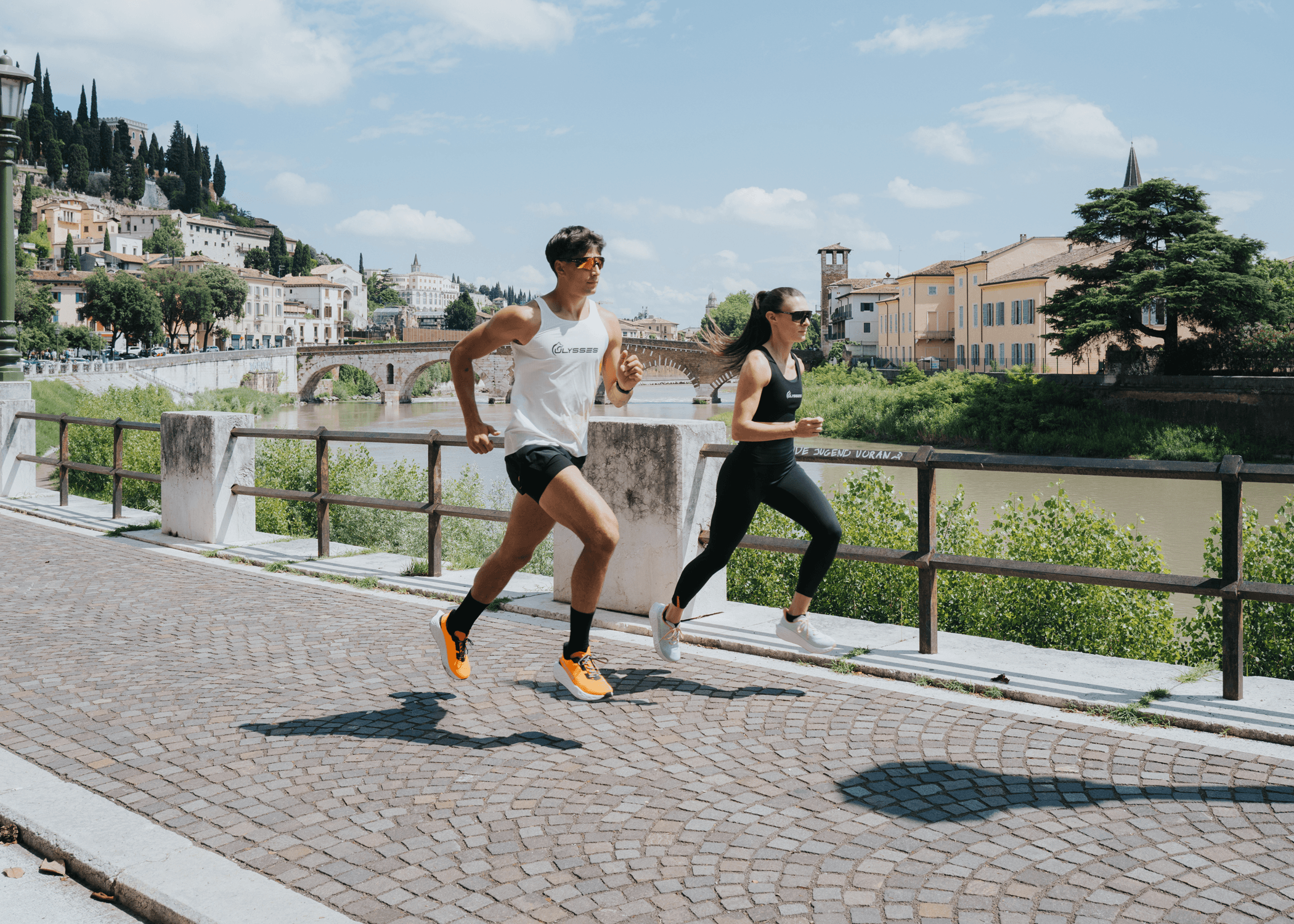 Man and woman running together along a riverside promenade in Verona, wearing Ulysses tank tops and shoes, with a historic stone bridge and hillside buildings in the background.