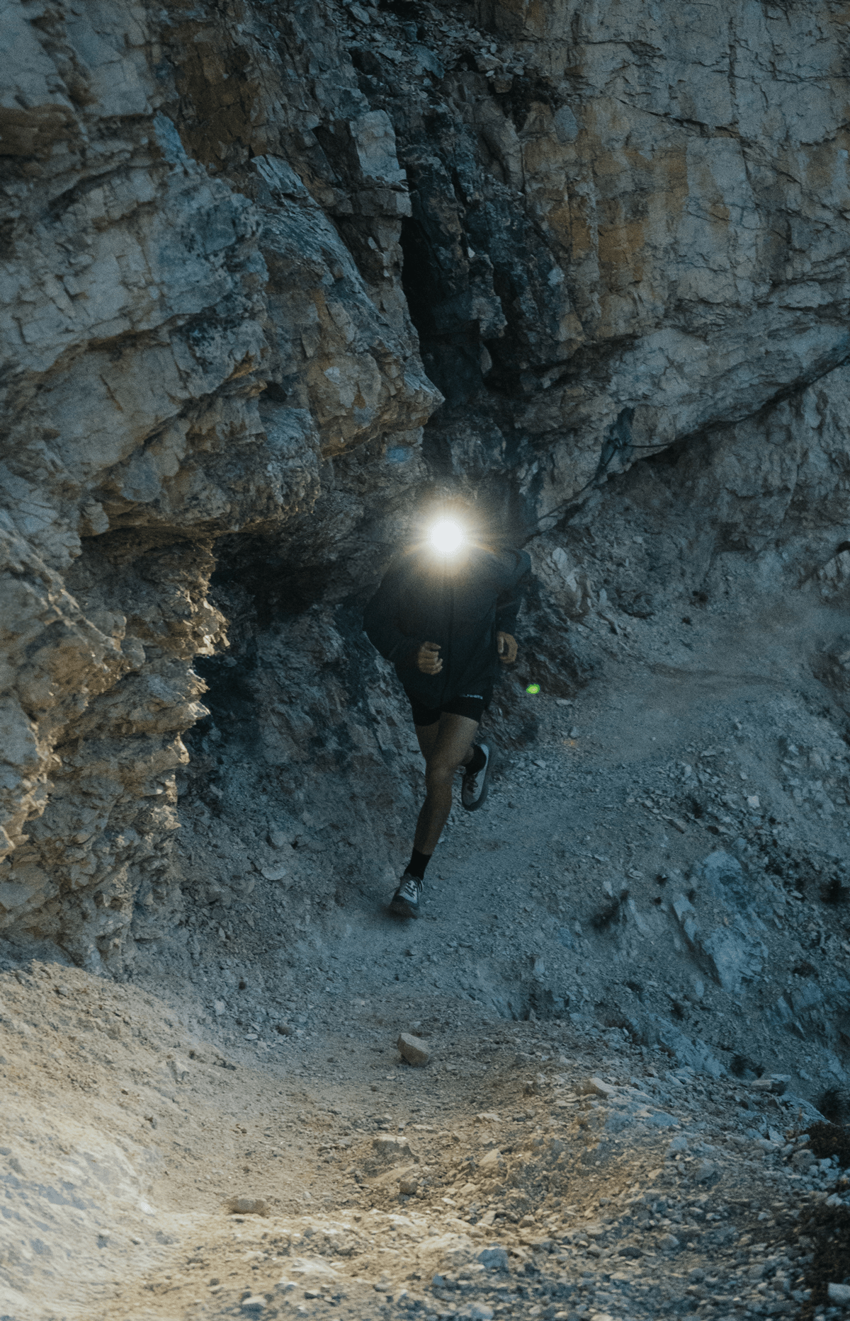 Trail runner with a headlamp navigating a narrow rocky gorge at dusk.