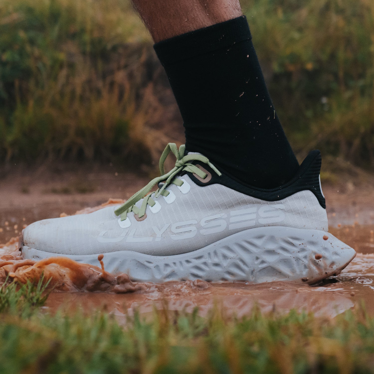 "First Impact Spoiler” – scarpa da corsa Ulysses bianca immersa nel fango durante una corsa off-road, che mostra la resistenza e la trazione della suola in un terreno naturale bagnato
