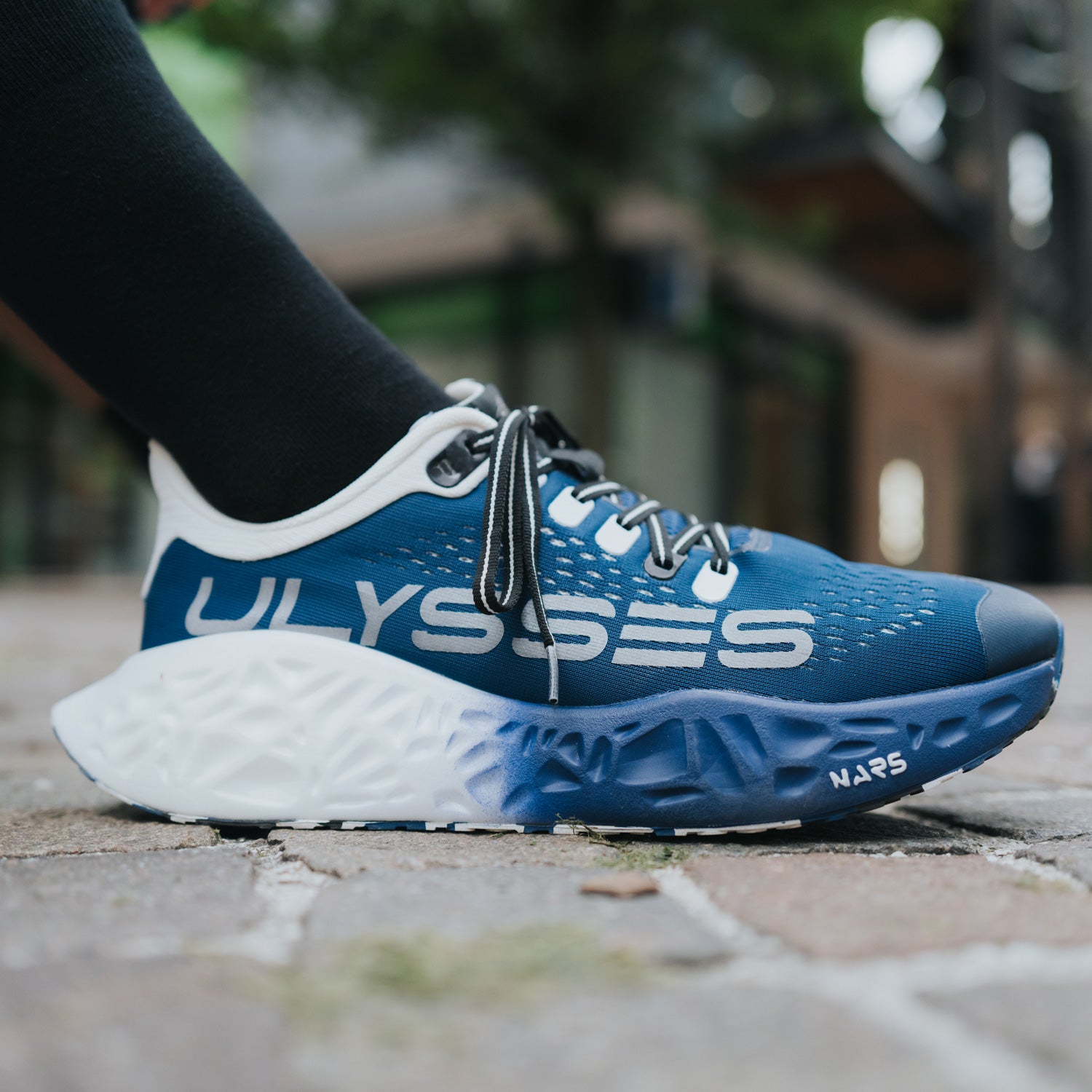 "G FAOM” – scarpa da corsa Ulysses blu indossata su una superficie urbana in pietra, che evidenzia il design moderno e la versatilità del modello in contesto cittadino