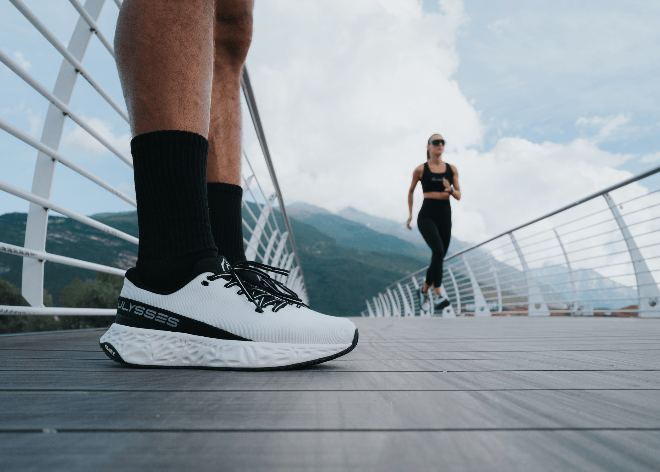 Close-up of a white and black Ulysses shoe on a man's foot on a bridge, with a woman running in the background against a mountain landscape.