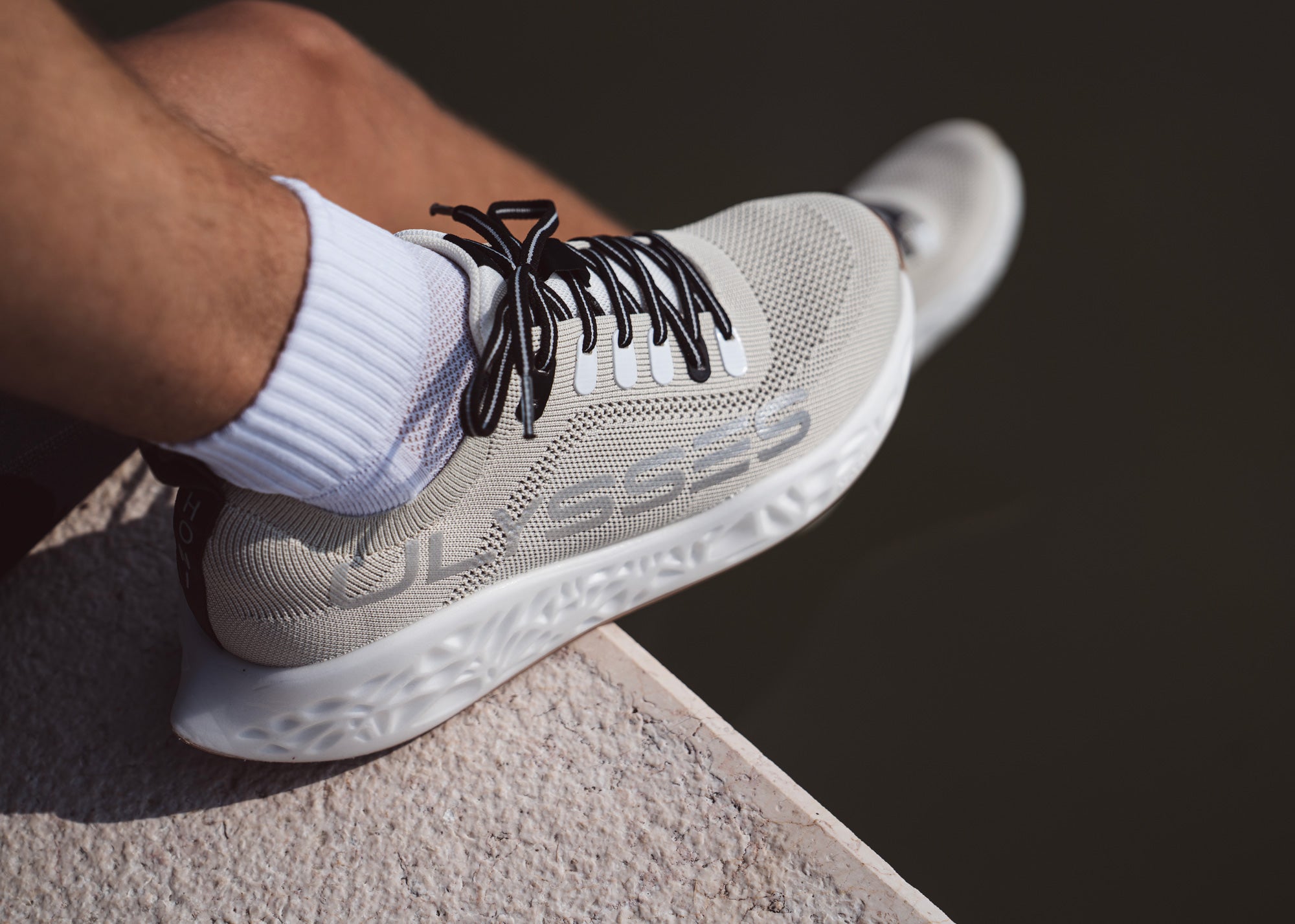 Close-up of the Ulysses Honi urban running shoe in light beige, worn by a person sitting on a ledge.