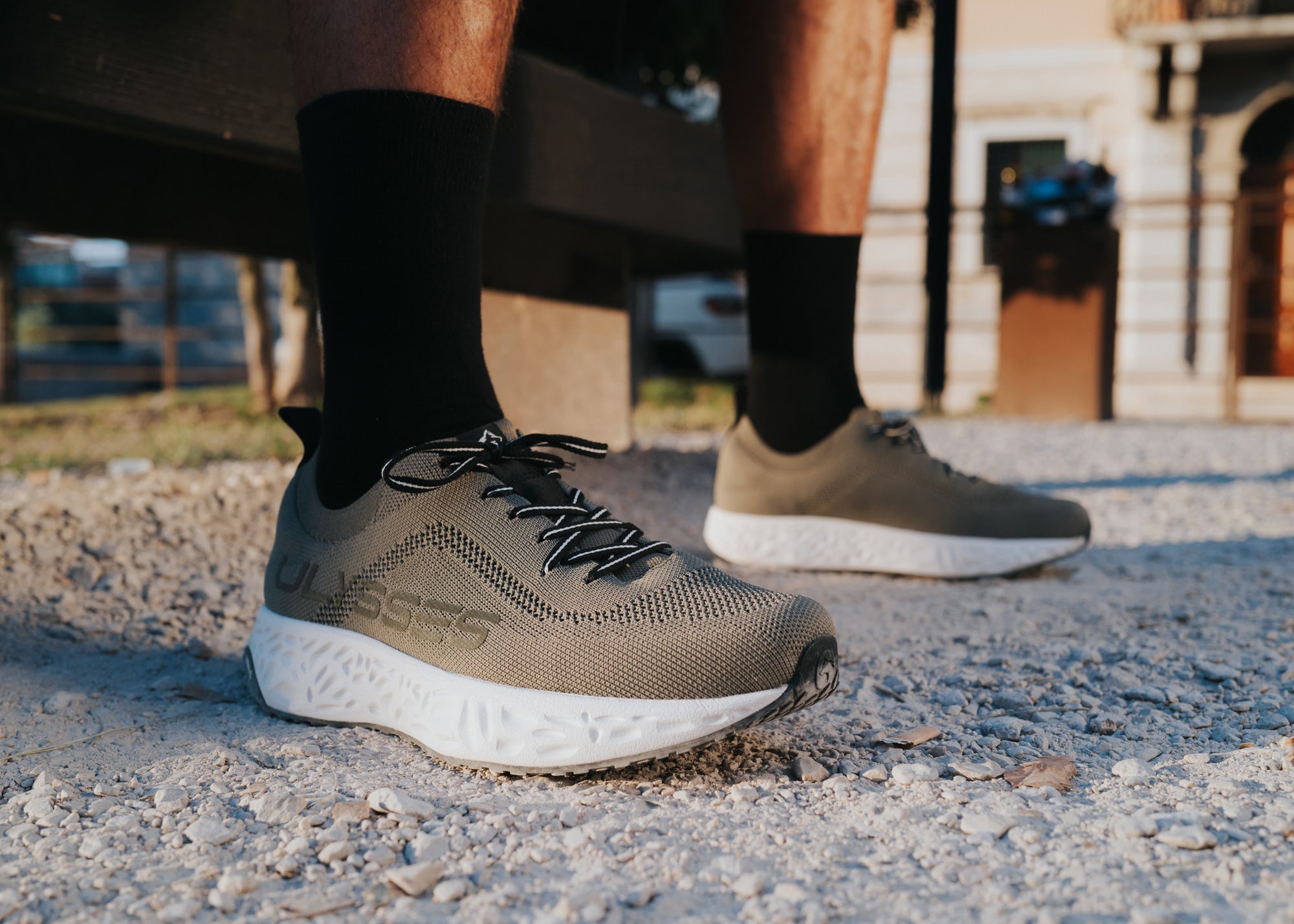 Close-up of olive green knit Ulysses shoes on a man's feet, standing on a gravel path near a building.