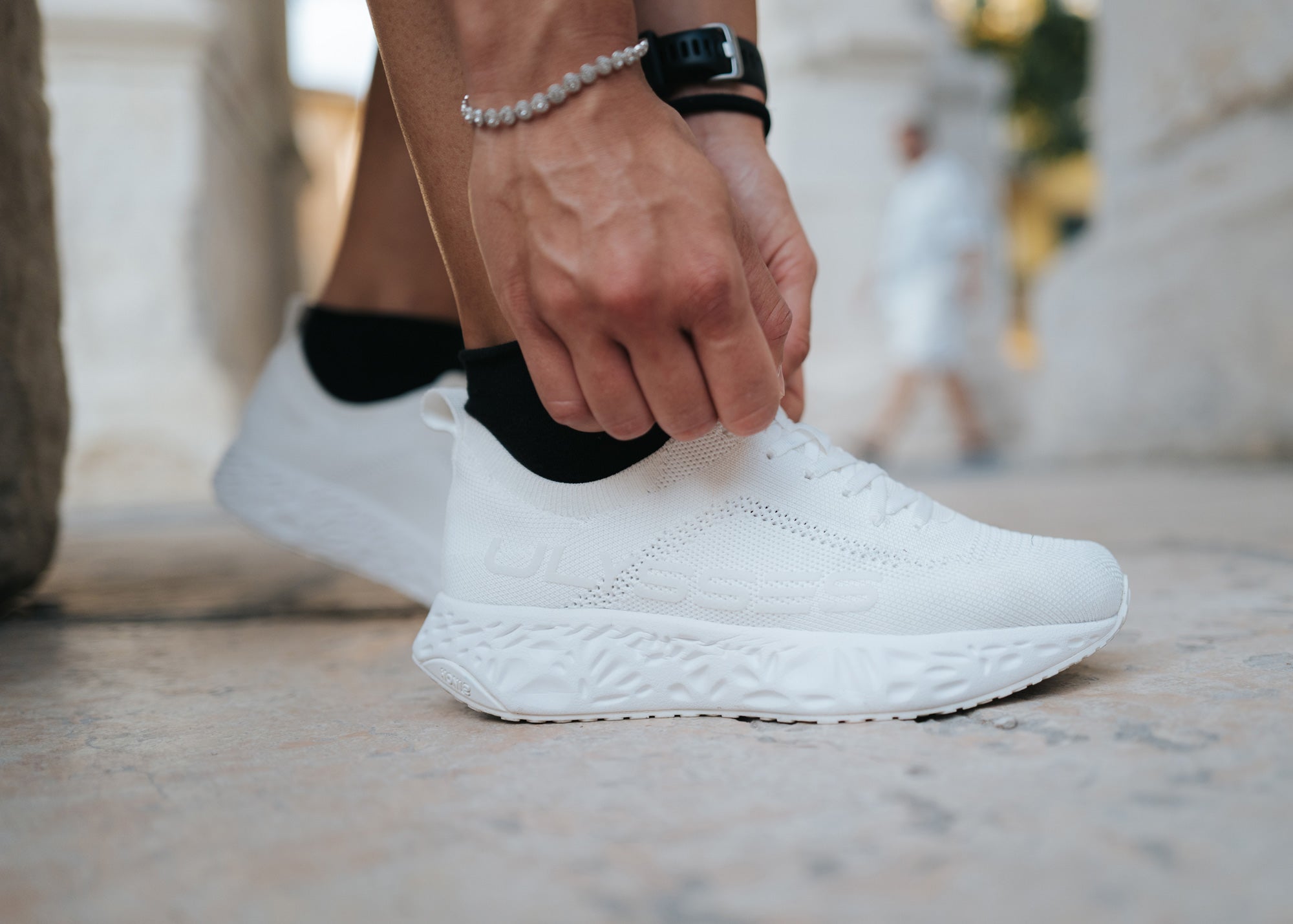 Close-up of a white knit Ulysses shoe as a hand adjusts the heel, with a bracelet and smartwatch visible on the wrist.