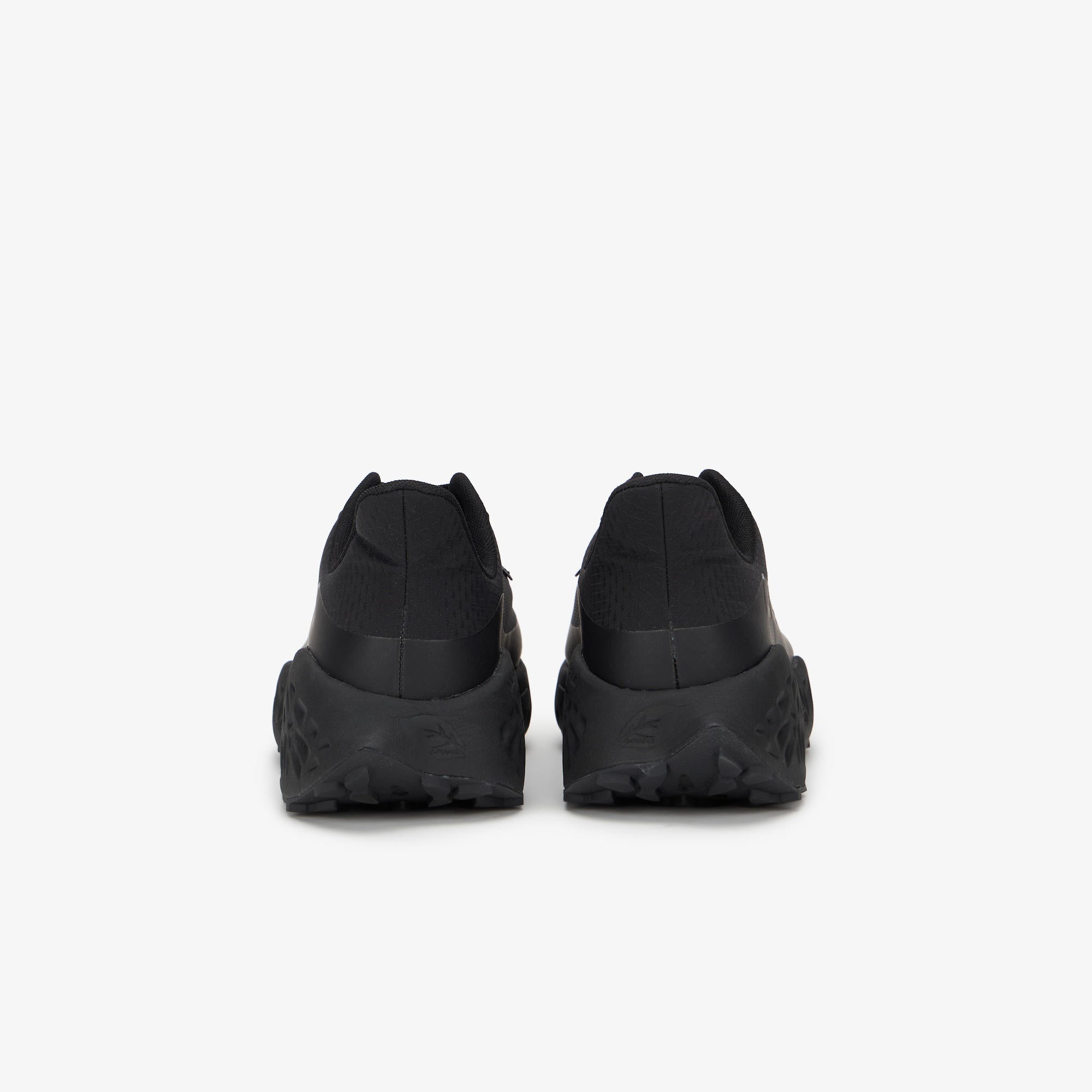 A pair of black low-top sneakers shown from the rear, featuring chunky black soles and a textured upper, set against a plain light gray background.