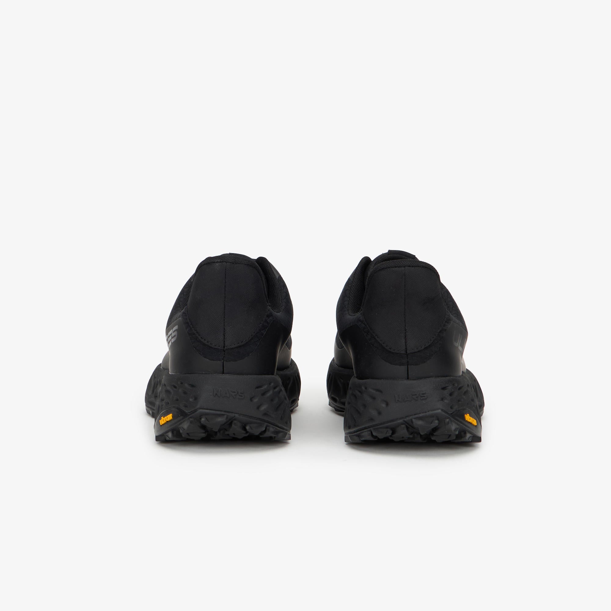 A pair of black athletic sneakers shown from the rear, featuring textured black soles and tiny orange Vibram logos on the outsoles, set against a plain light gray background.