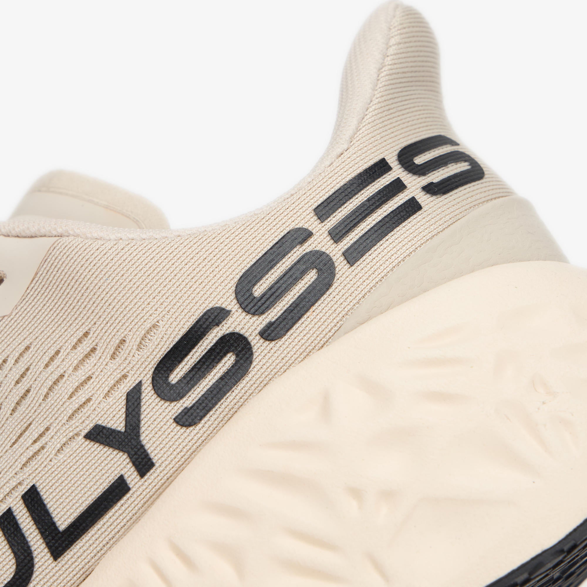 Close-up of a beige athletic shoe showing the heel area with a black 'ULYSSES' logo printed across the side and a textured beige sole.