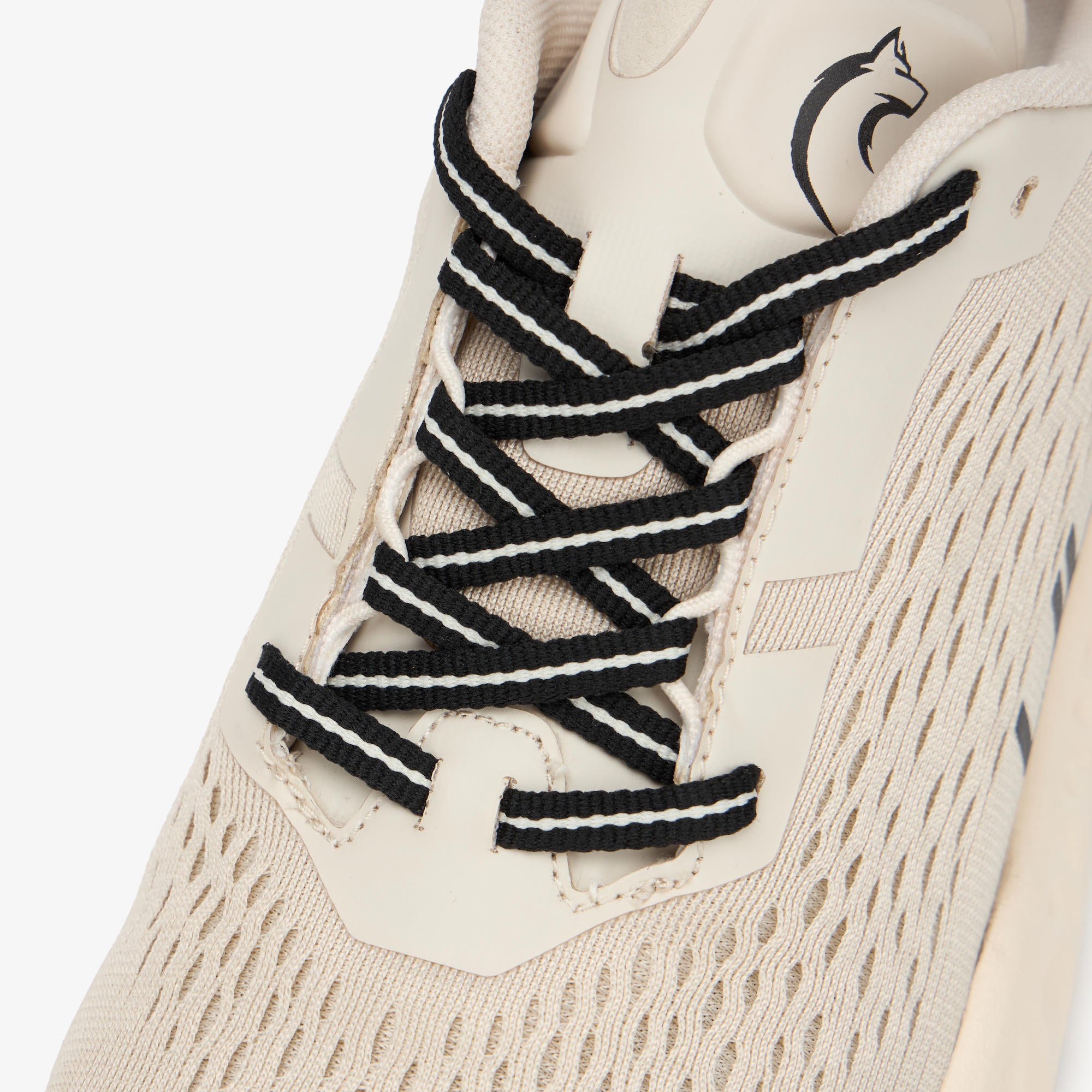 Close-up of a beige athletic sneaker with black and white striped laces, showing the textured mesh upper and a black logo on the tongue.