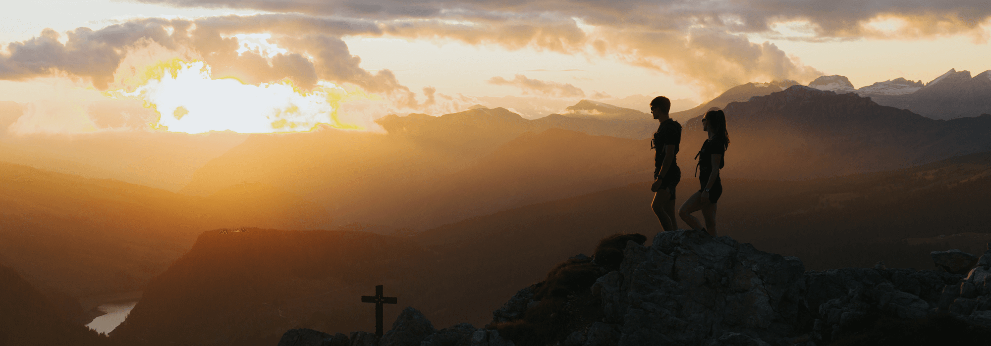 Two trail runners standing on a rocky mountain ridge at sunset, overlooking layers of golden-lit peaks, symbolising endurance and outdoor exploration.