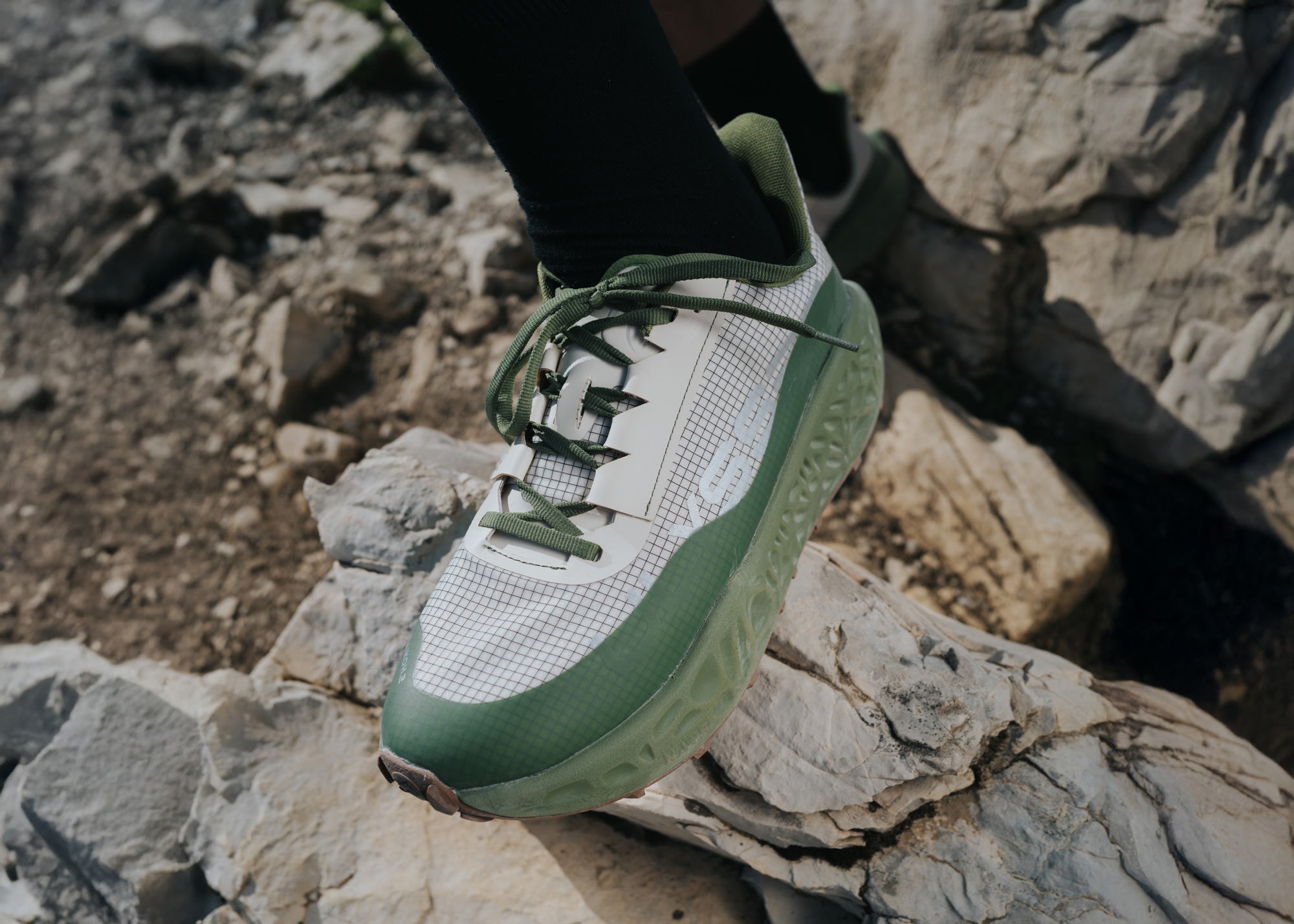 Close-up of a white and green Ulysses trail shoe on a man's foot, positioned on a rocky outcrop.
