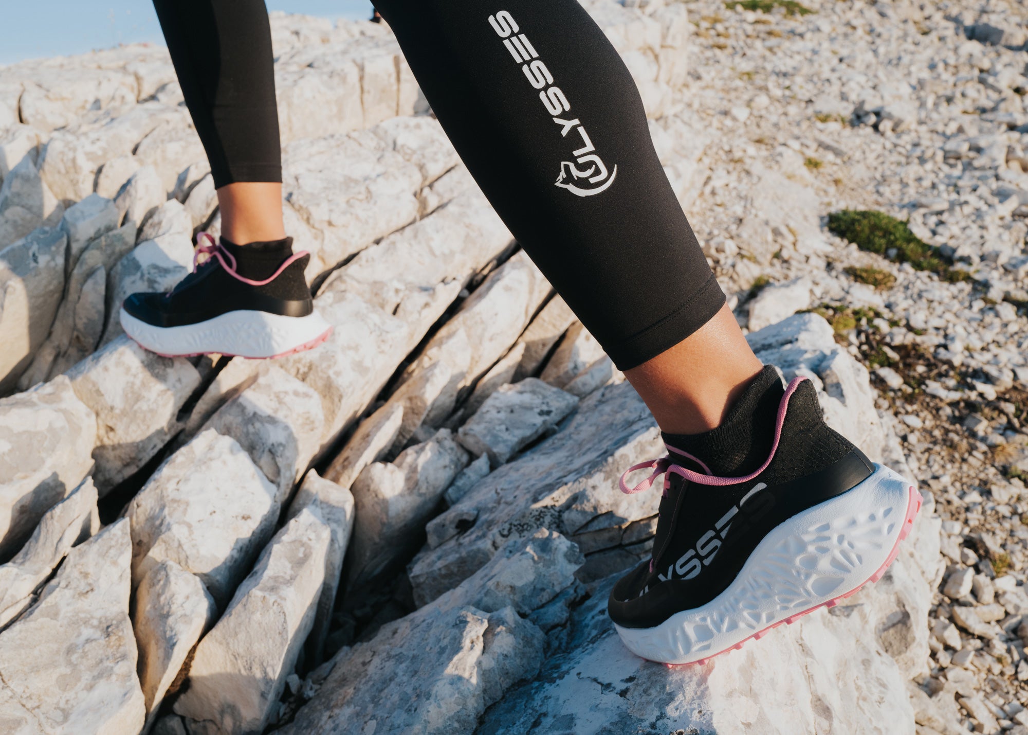 Close-up of black Ulysses trail shoes with pink laces on a woman's feet, stepping across rocky terrain, with "ULYSSES" branding visible on her leggings.