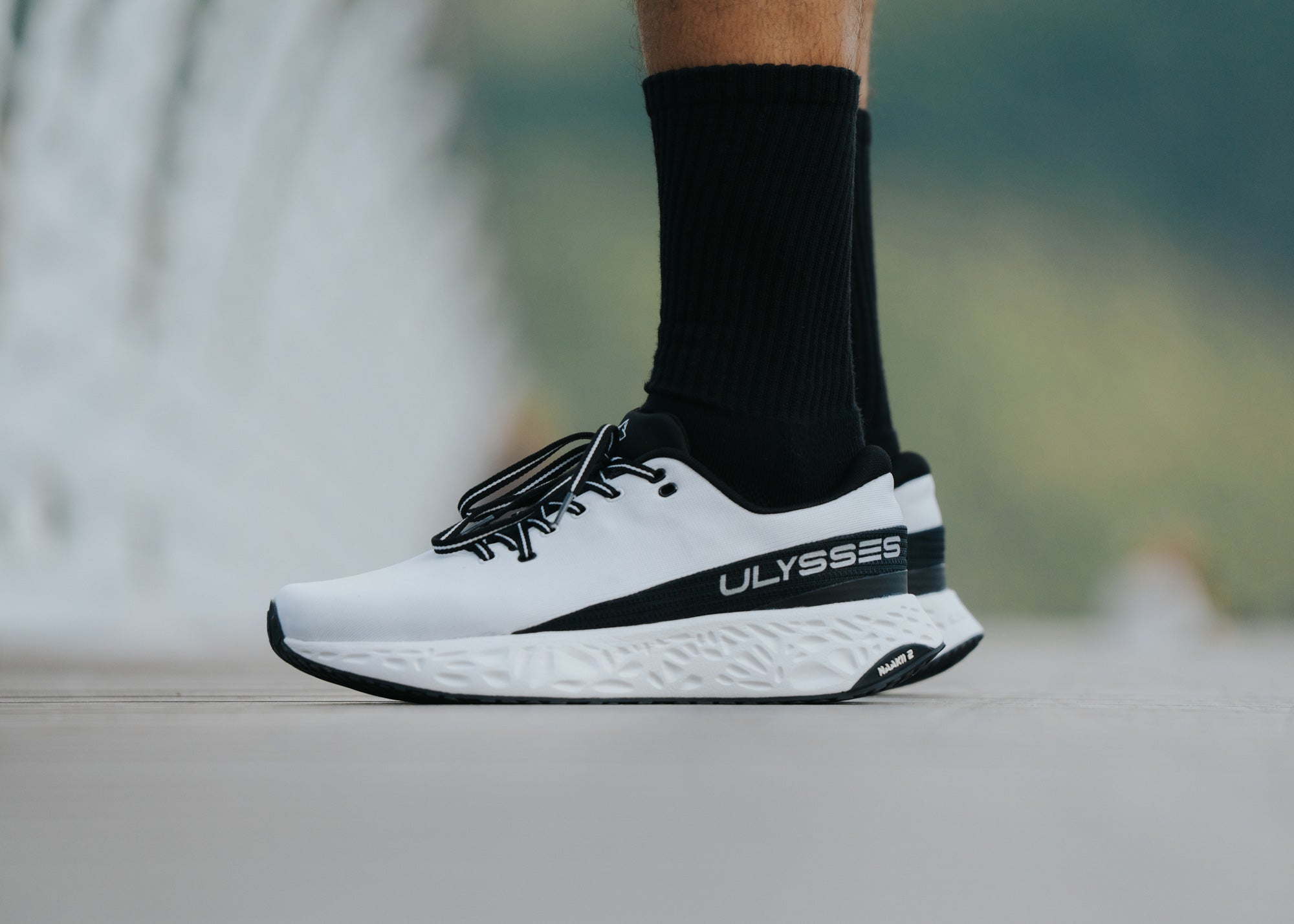 Side profile close-up of a white and black Ulysses running shoe on a man's foot, showing the "ULYSSES" branding and Naakii 2 outsole.