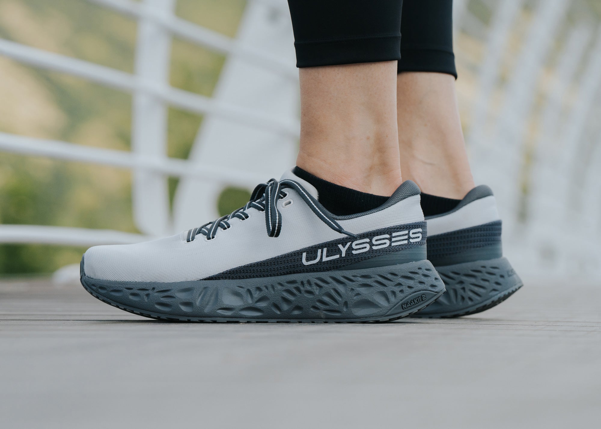 Side profile close-up of a light gray and charcoal Ulysses running shoe on a woman's foot, standing on a bridge with a railing visible in the background.