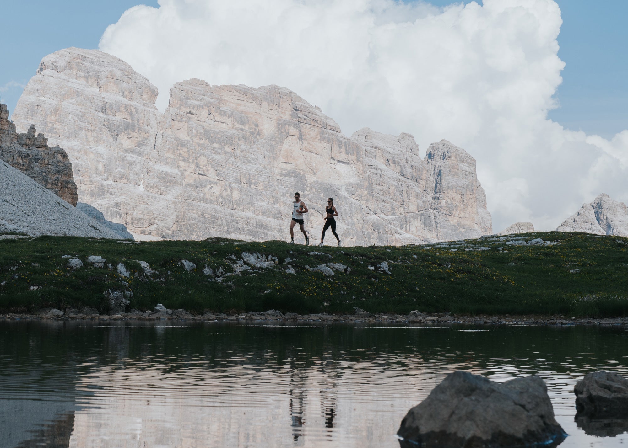 Man and woman trail running along a grassy ridge above a mountain lake, with dramatic Dolomite peaks and clouds in the background.