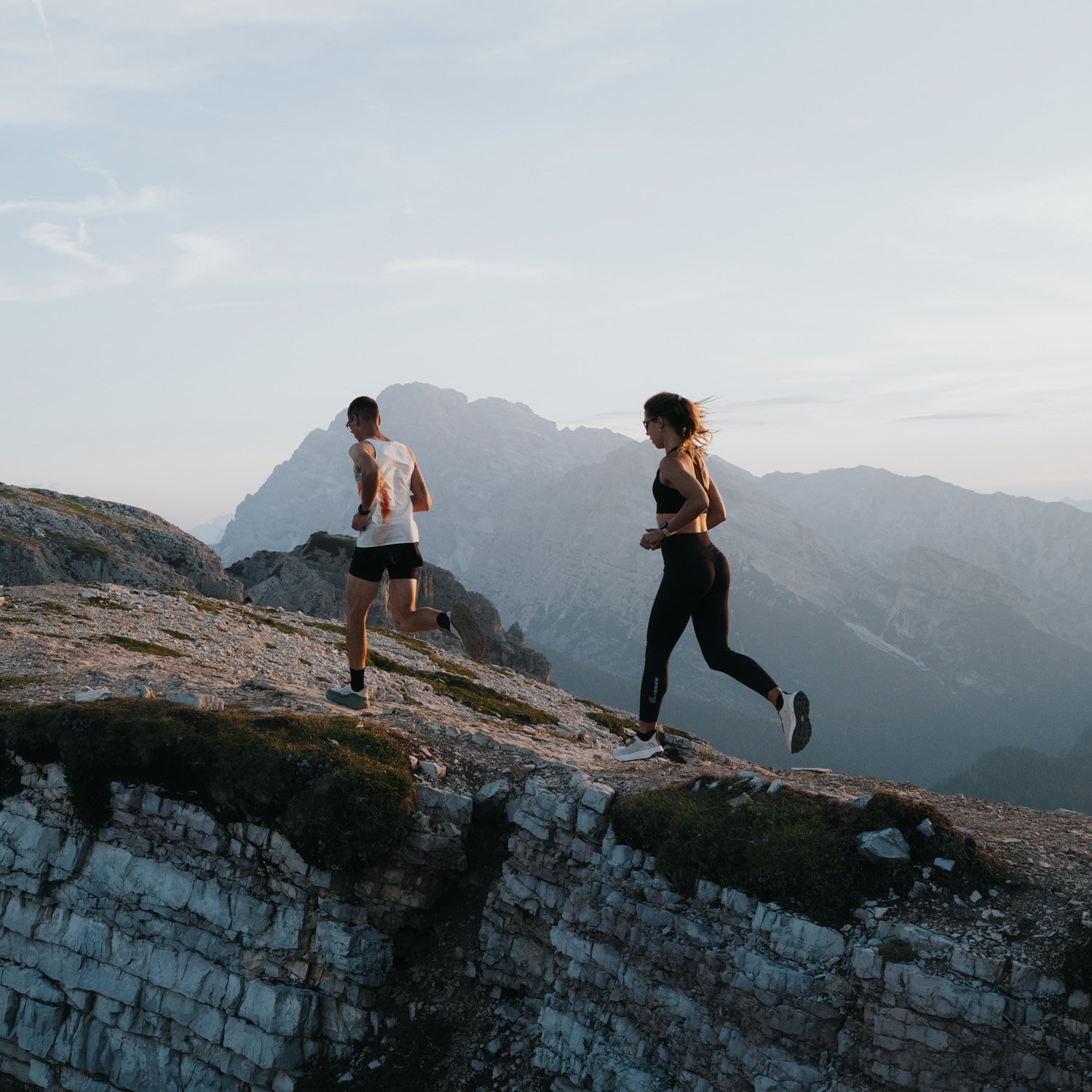 Two runners jogging on a rocky mountain ridge with hazy mountains in the background under a pale sky.