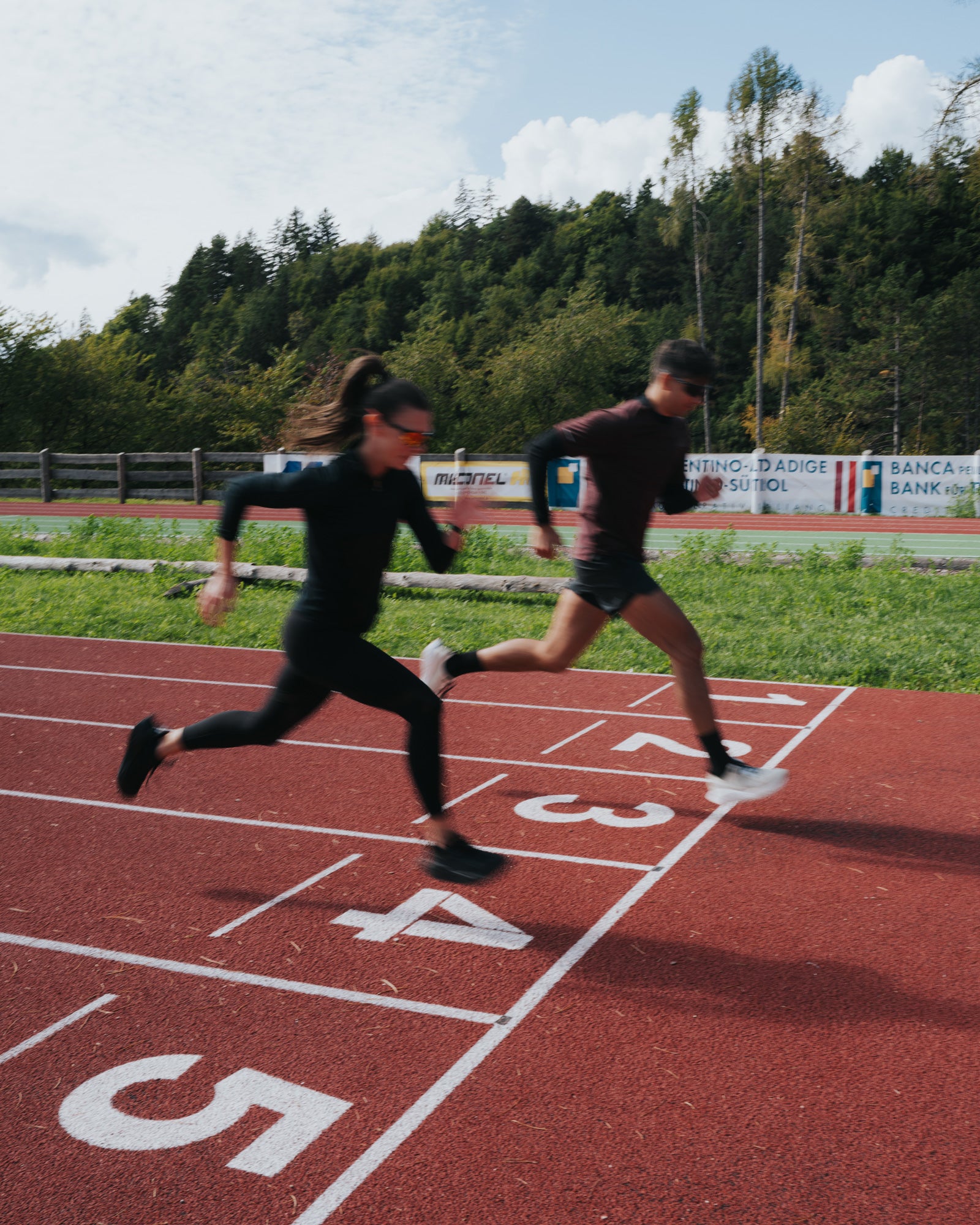 Two athletes sprint side by side on a red running track, wearing Ulysses Road running shoes, captured in motion as they cross the finish line with determination.