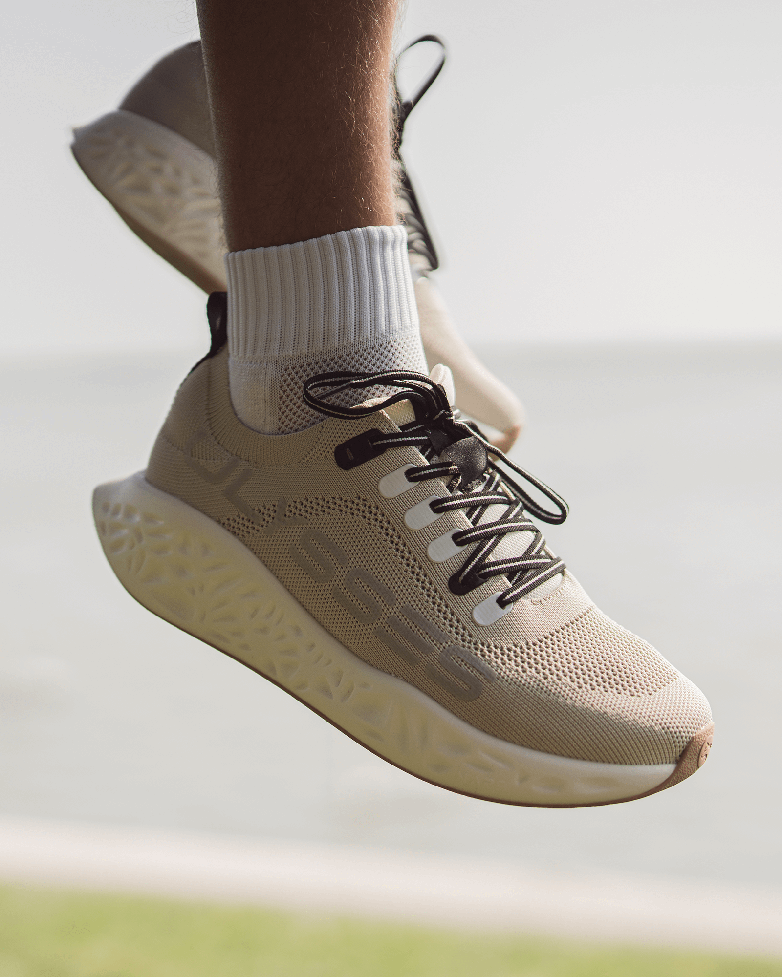 Close-up of beige Ulysses running shoes mid-air, highlighting the textured midsole design and breathable upper material.