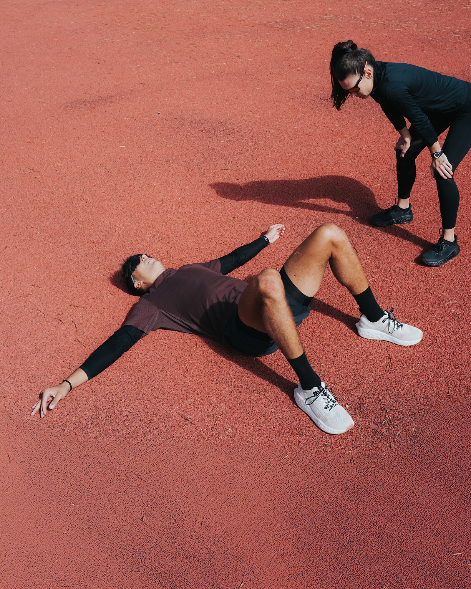 Runner lying exhausted on a red athletic track while another runner stands nearby, both wearing Ulysses running shoes during an intense training session.