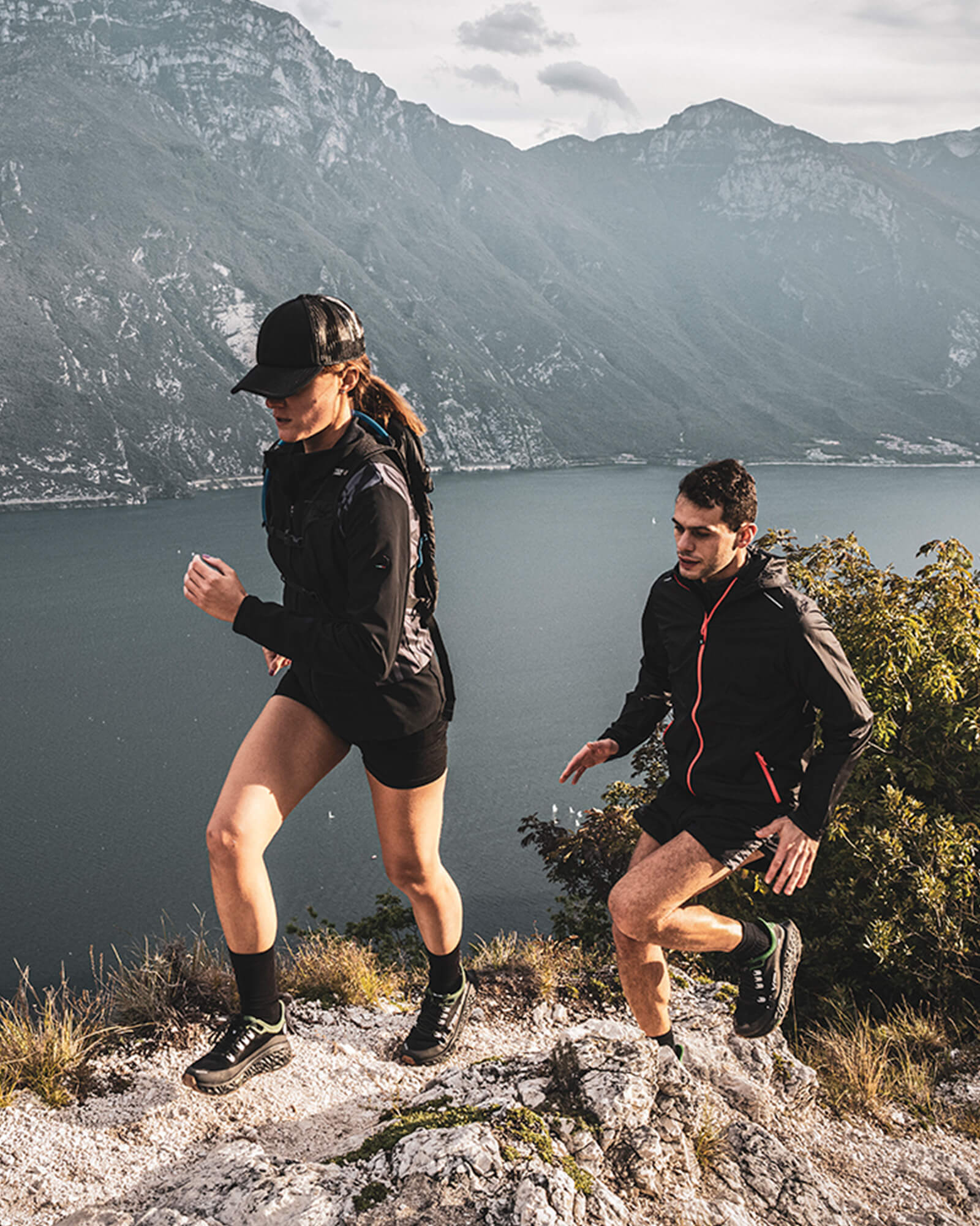 Two trail runners climbing a rocky path above a lake, surrounded by steep mountains, wearing Ulysses trail shoes.