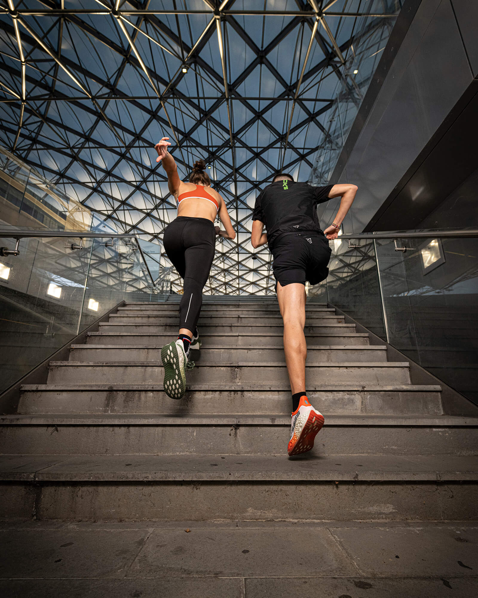A man and a woman running up concrete stairs inside a modern glass-covered structure, wearing Ulysses running shoes.