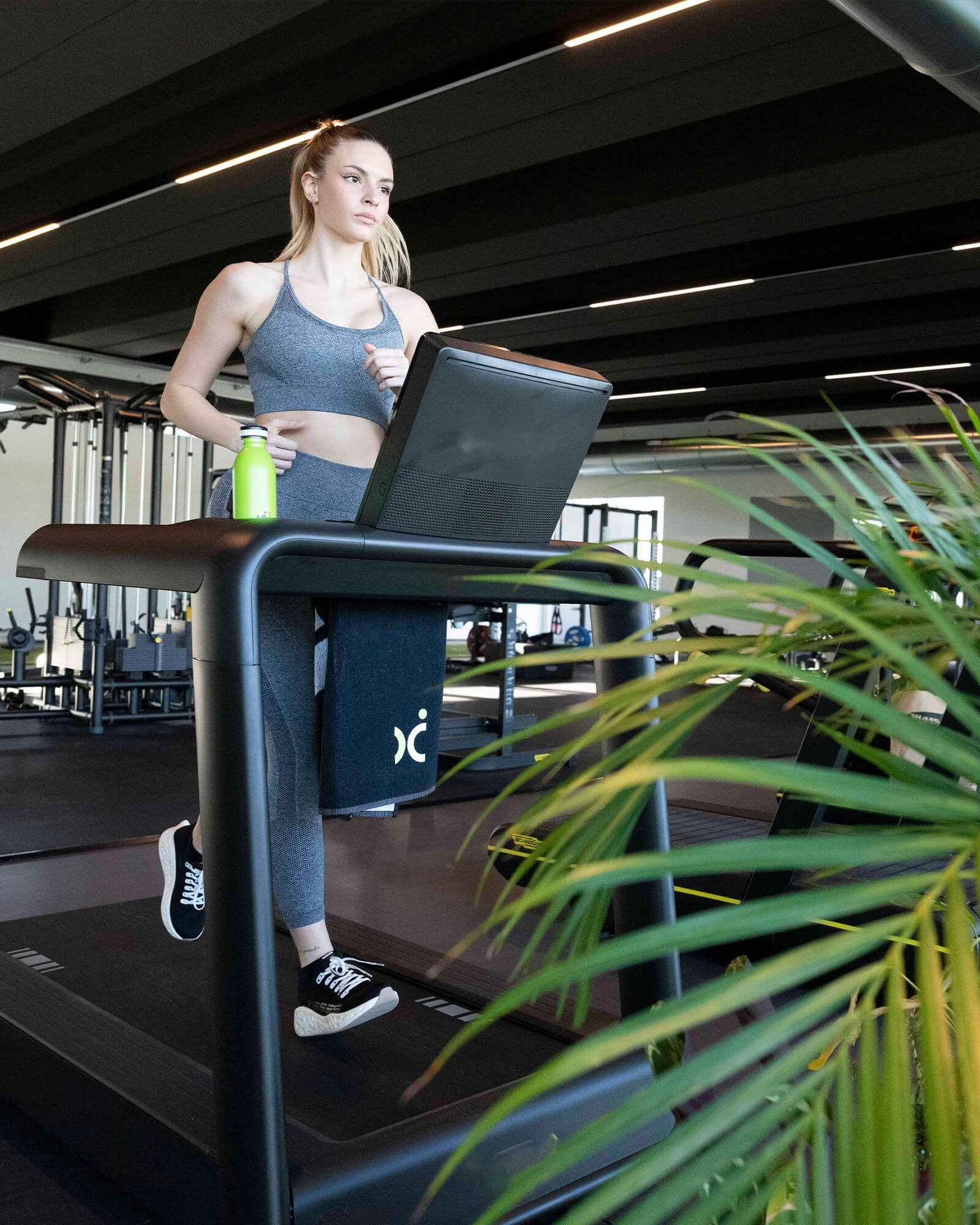 A woman running indoors on a treadmill in a gym, wearing black Ulysses running shoes and grey sportswear.