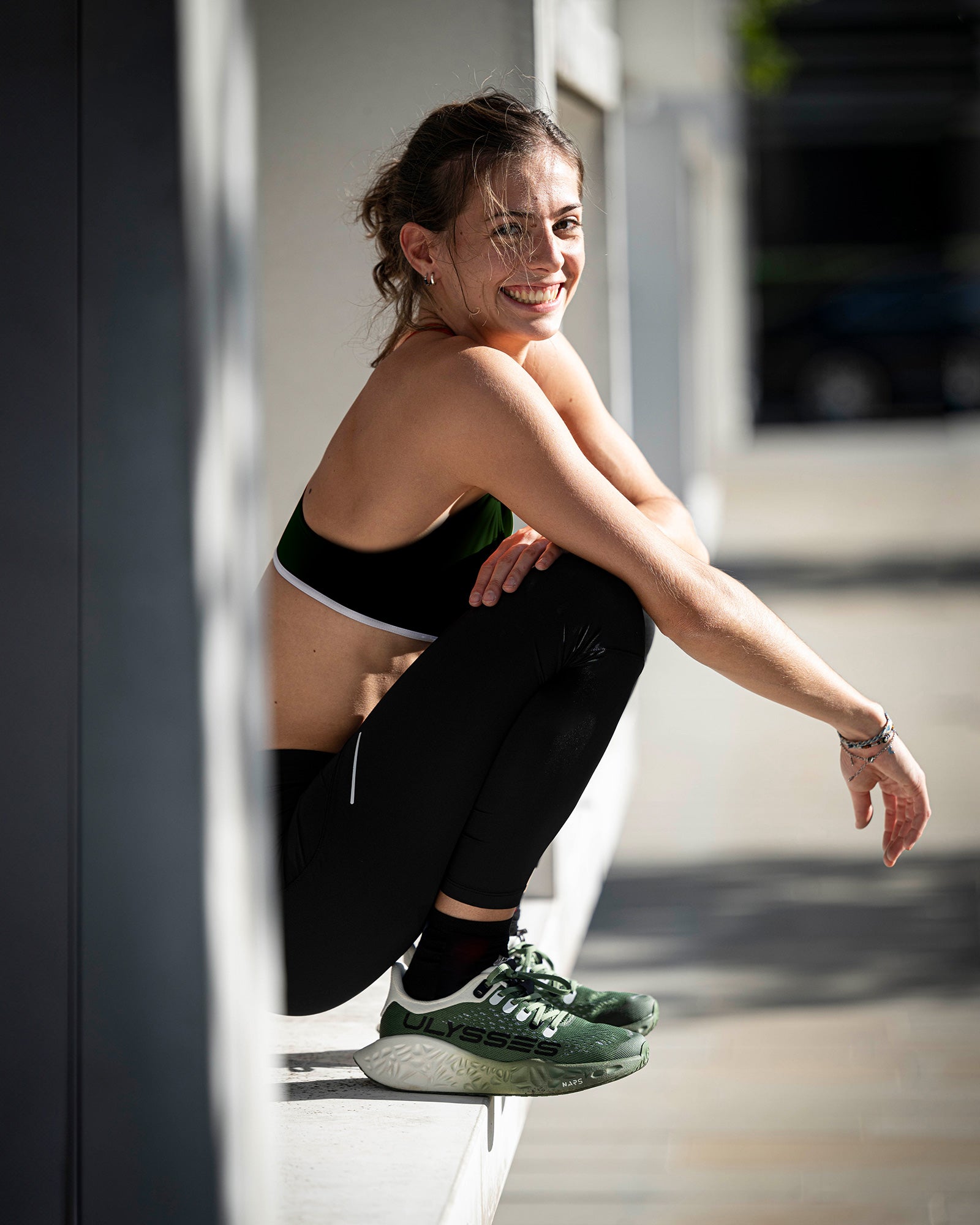 A smiling woman sitting on an outdoor ledge after a workout, wearing green Ulysses running shoes.