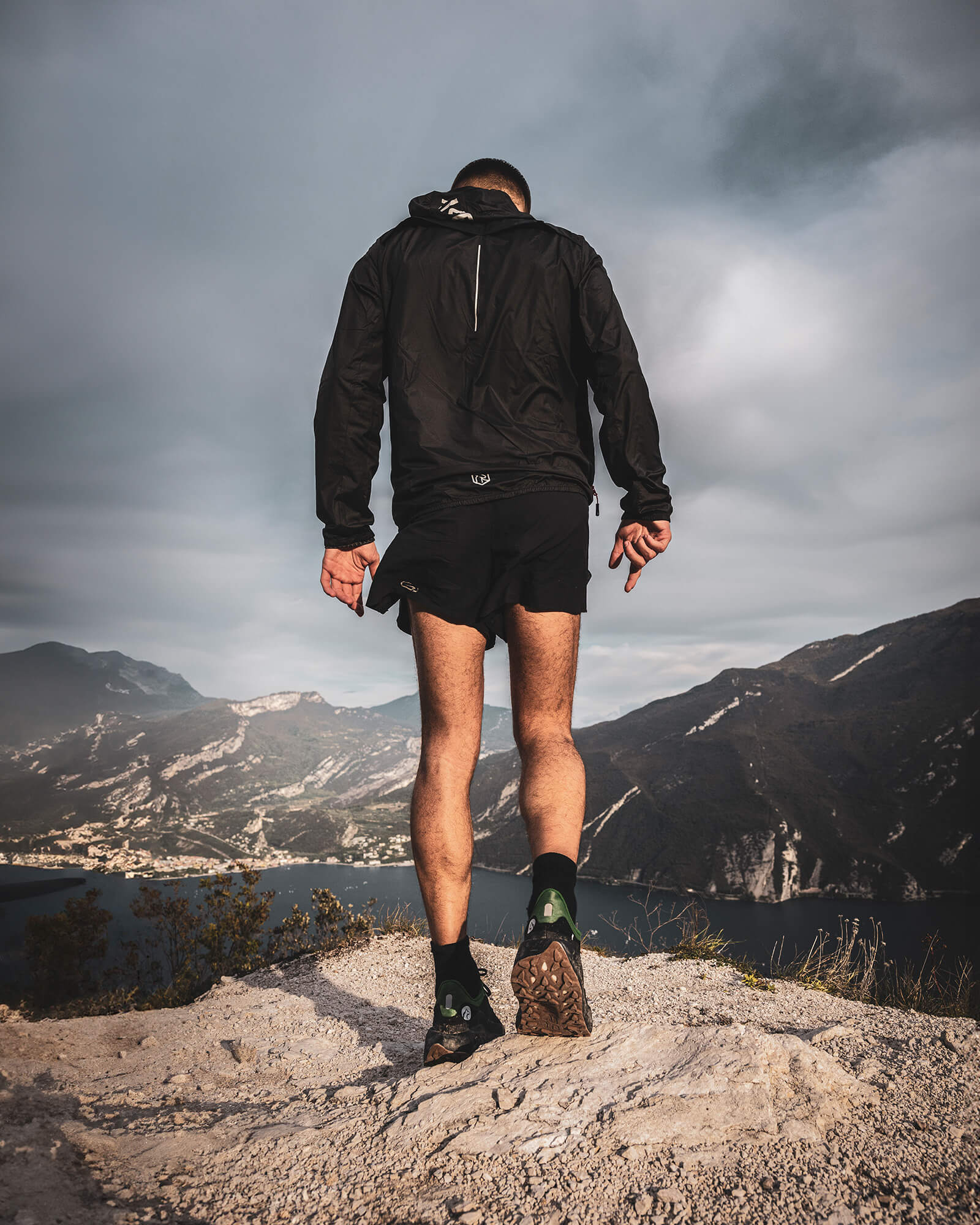 A trail runner standing on a rocky peak overlooking a lake and mountains, wearing black Ulysses trail shoes.