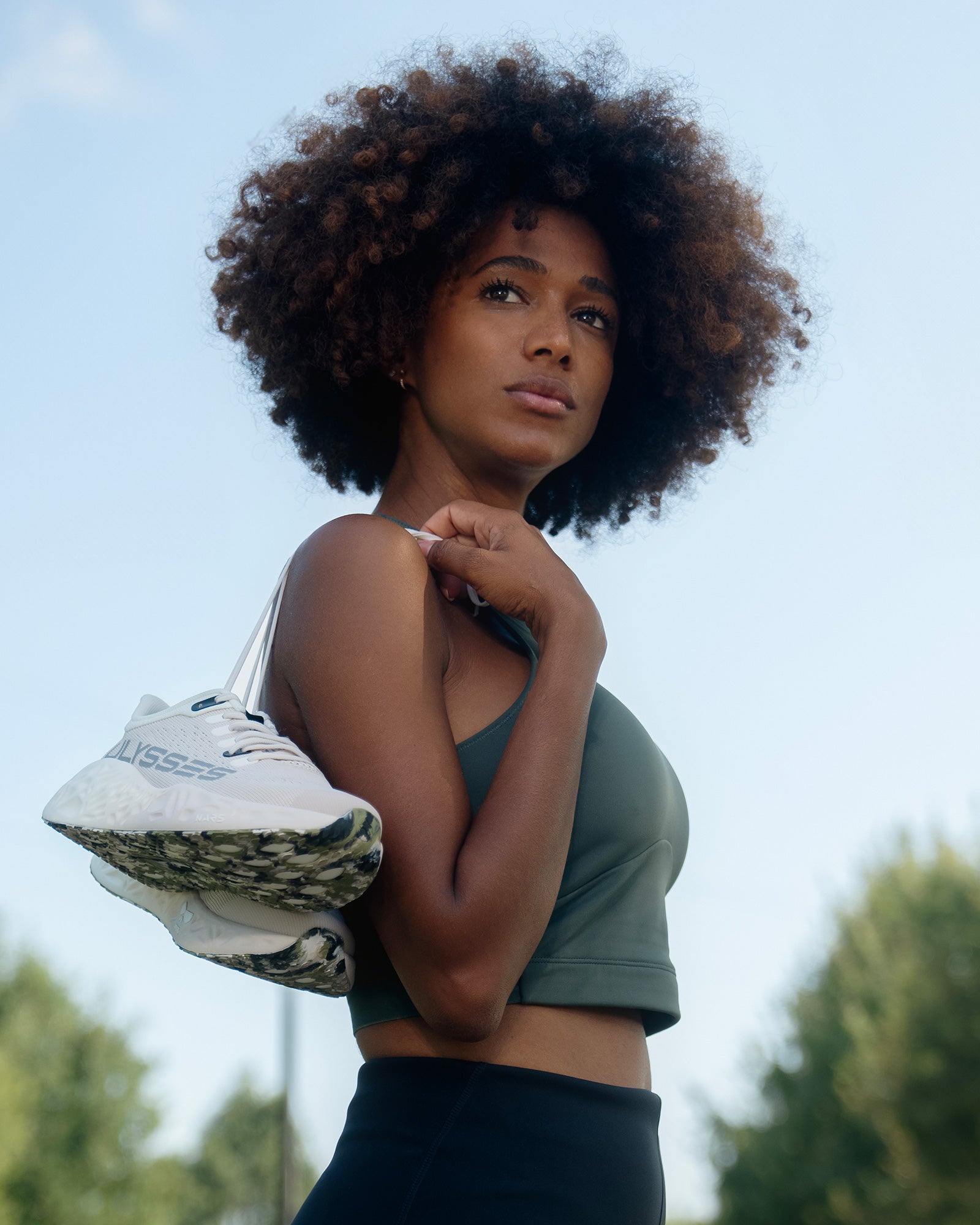 A woman outdoors holding white Ulysses running shoes over her shoulder, looking into the distance under a clear sky.