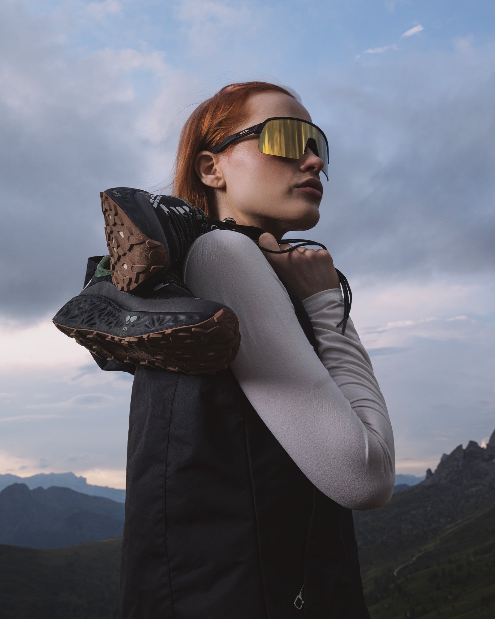 A woman standing outdoors in the mountains at dusk, carrying black Ulysses trail shoes over her shoulder and wearing reflective sunglasses.