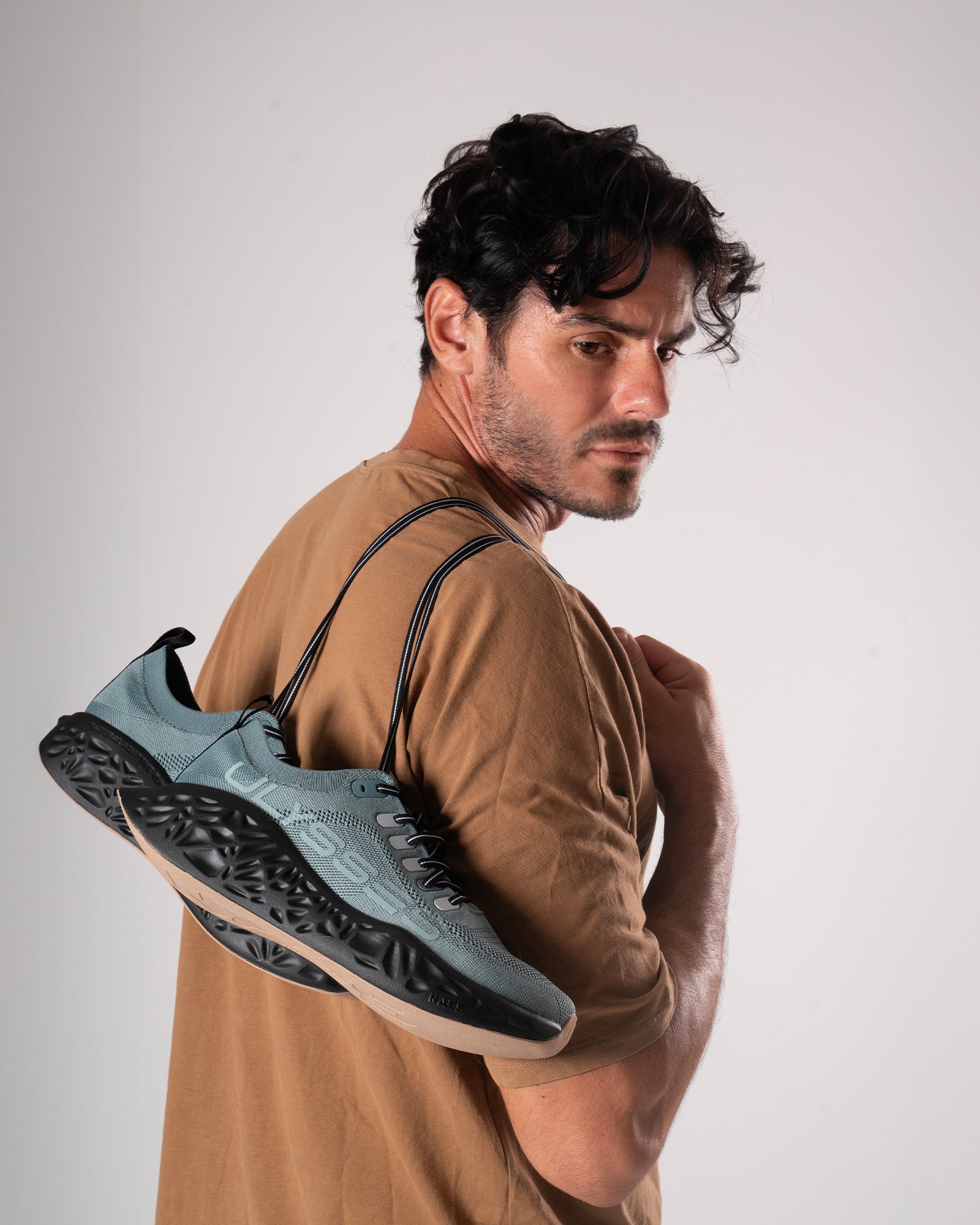 A man posing in a studio with a pair of teal Ulysses running shoes hanging over his shoulder.