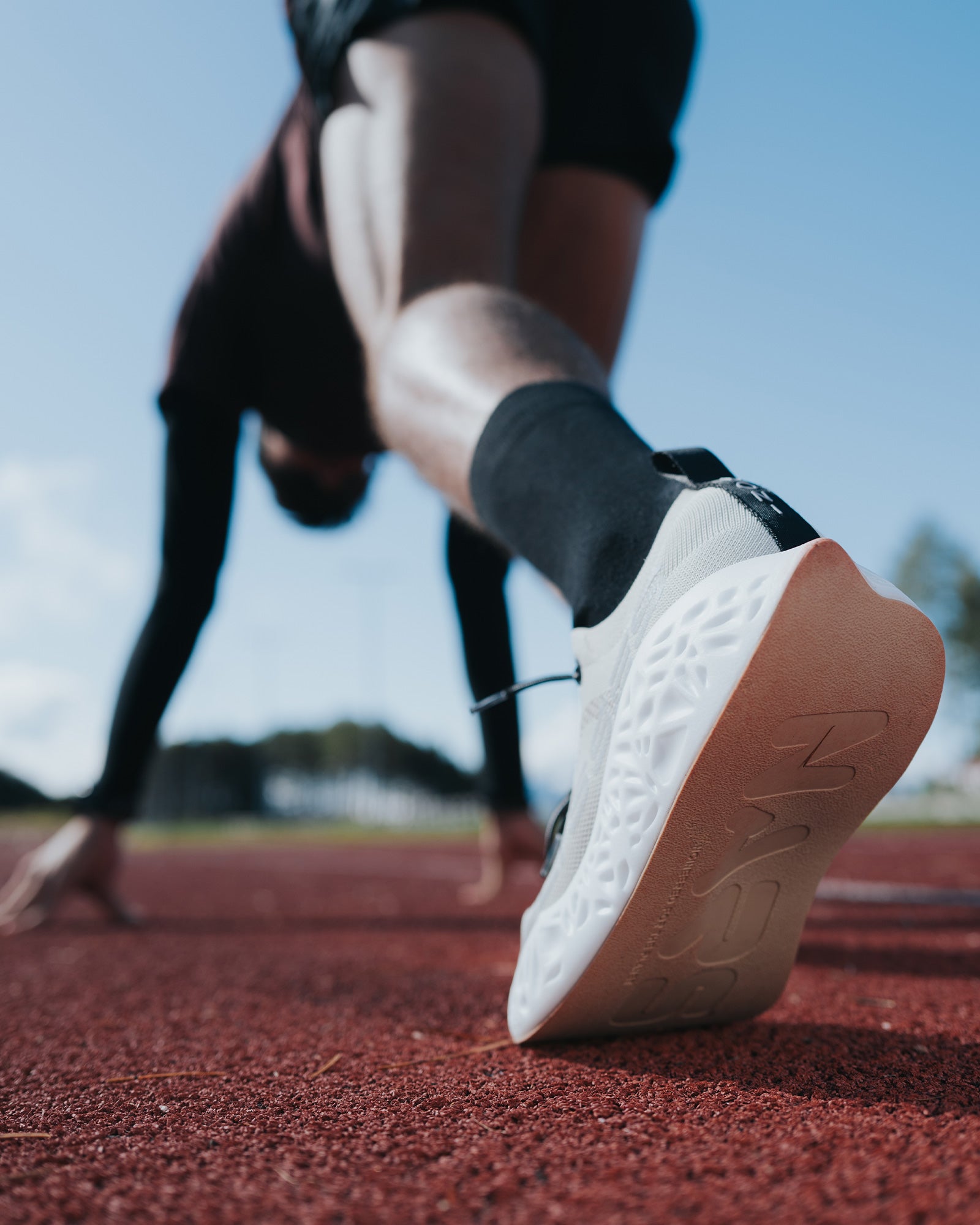 A runner in the starting position on a track, captured from a low angle with a close-up of the Ulysses running shoe sole.