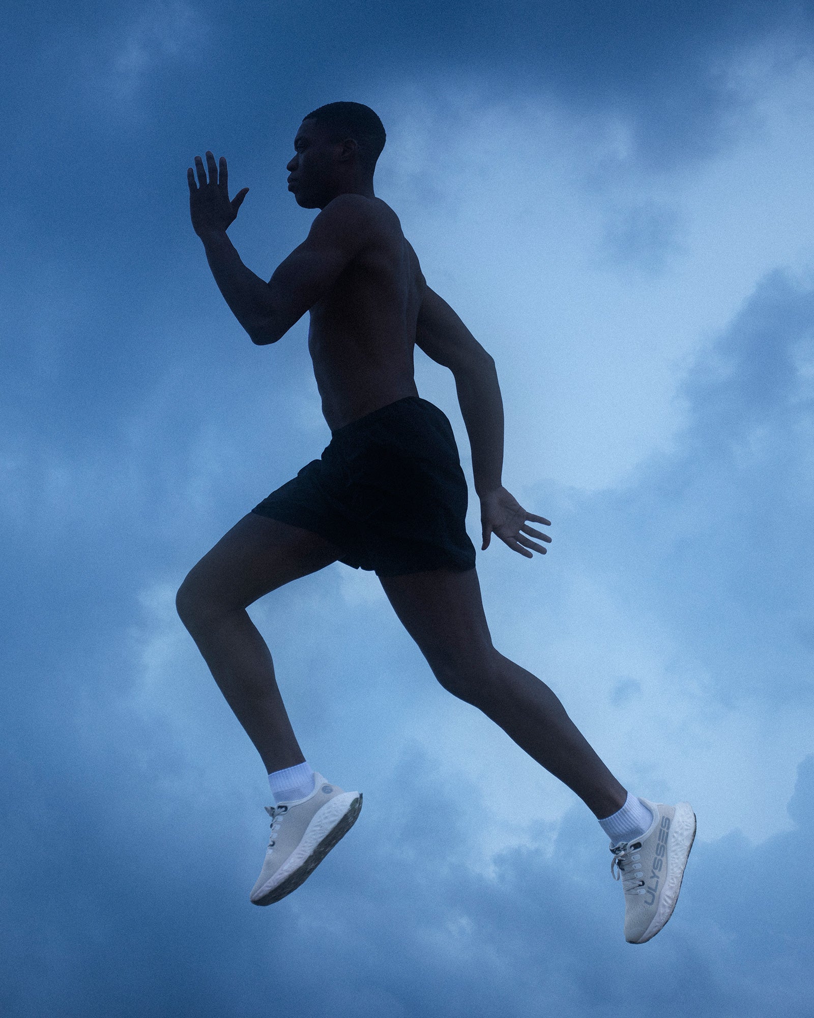 A shirtless athlete running against a dramatic cloudy sky, wearing white Ulysses running shoes.