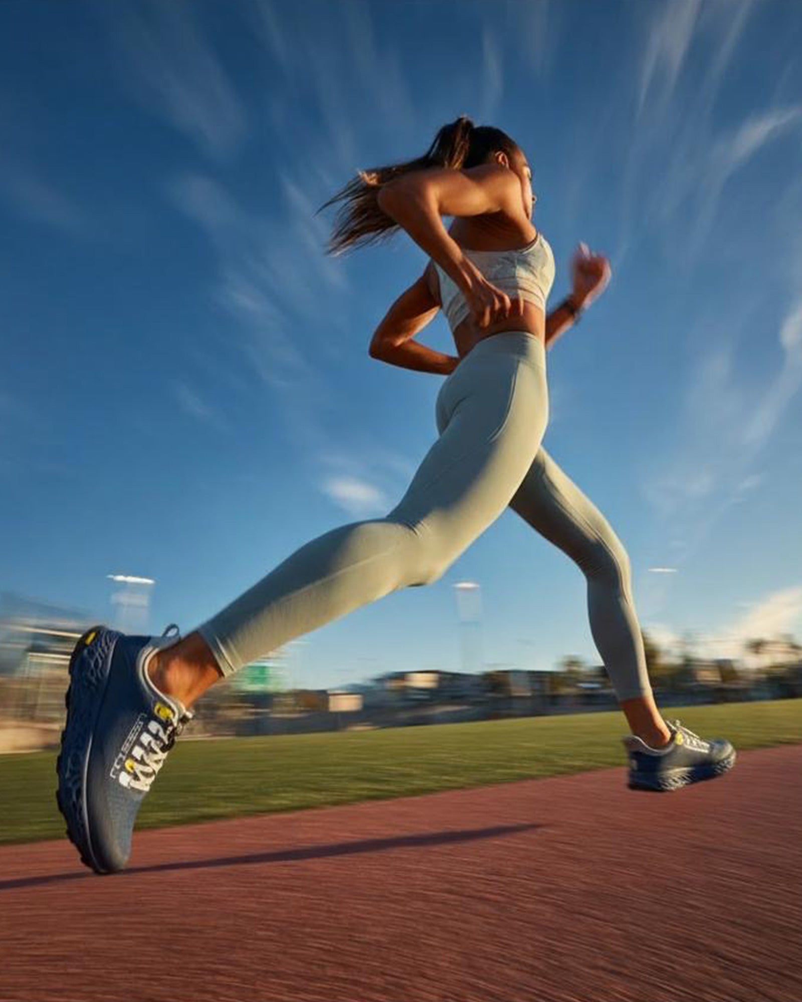 A woman sprinting on an outdoor track at sunset, wearing blue Ulysses running shoes.