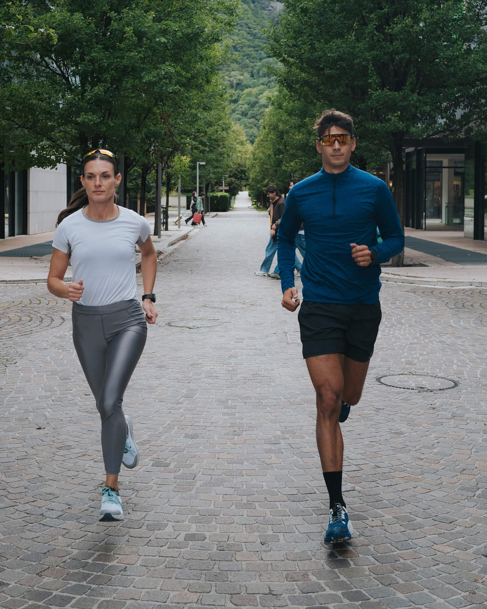 Two runners training together on a cobblestone street lined with trees, wearing Ulysses running shoes during an urban running session.
