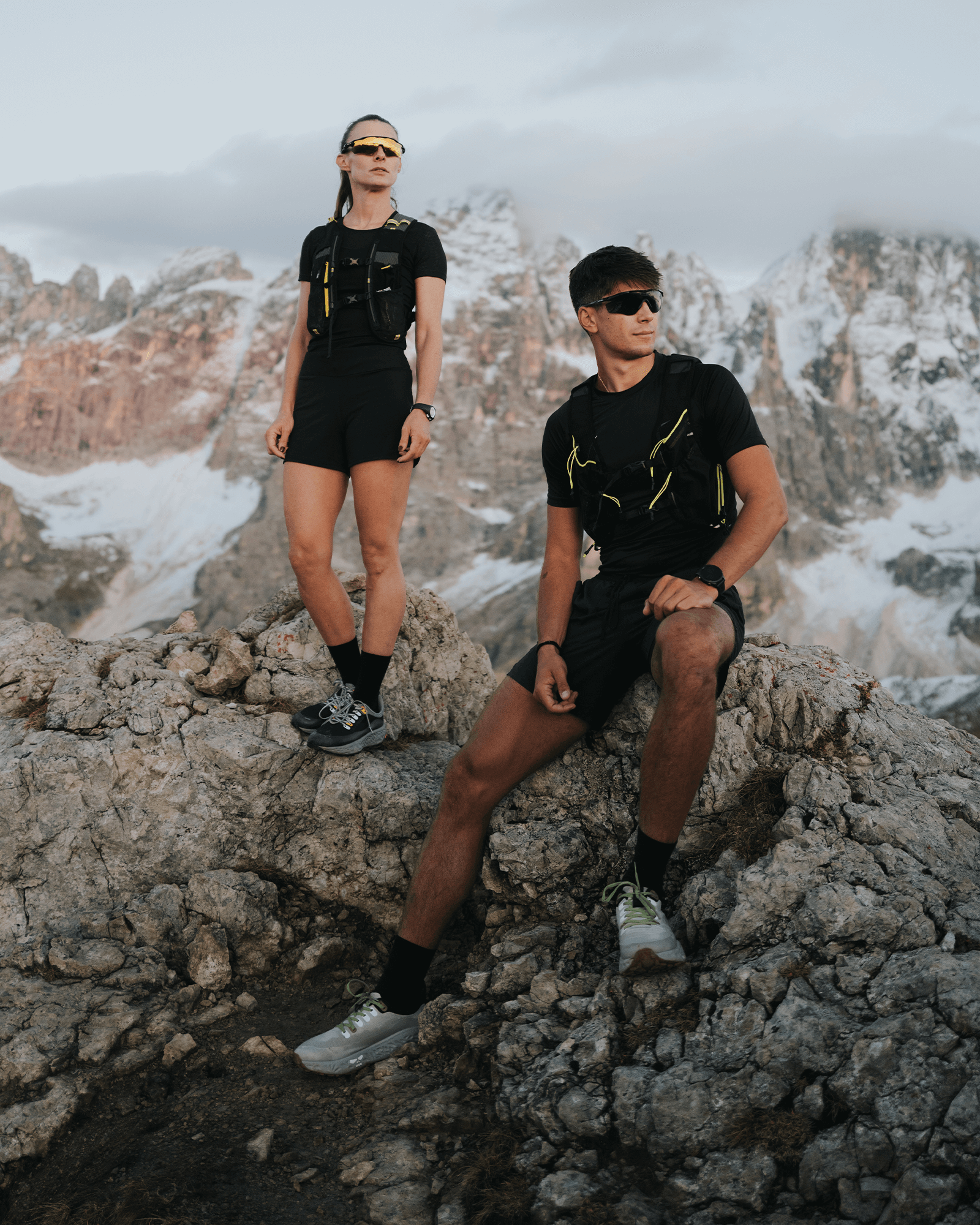 Two athletes standing on rugged mountain rocks with snowy peaks in the background, wearing Ulysses trail running gear and shoes.