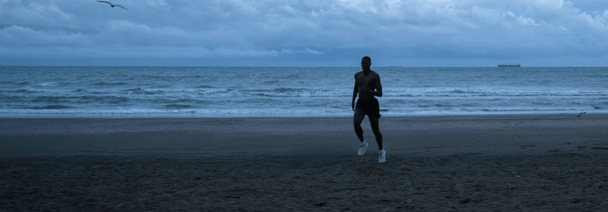 Solo runner jogging on a beach at dawn, with waves and a ship in the distance, wearing white Ulysses running shoes.