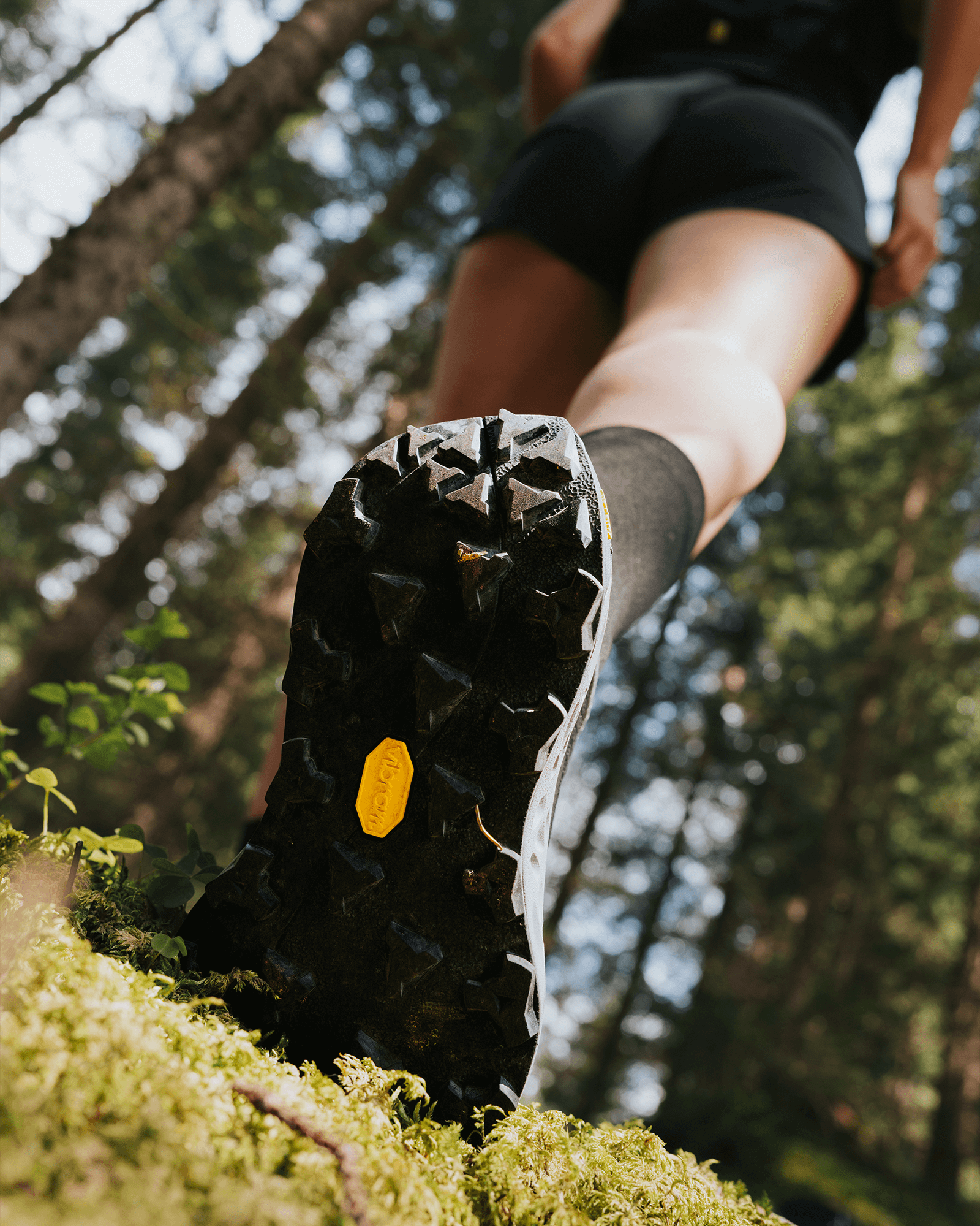 Bottom-up view of a trail runner’s shoe outsole with deep Vibram lugs, stepping on moss in a forest environment.