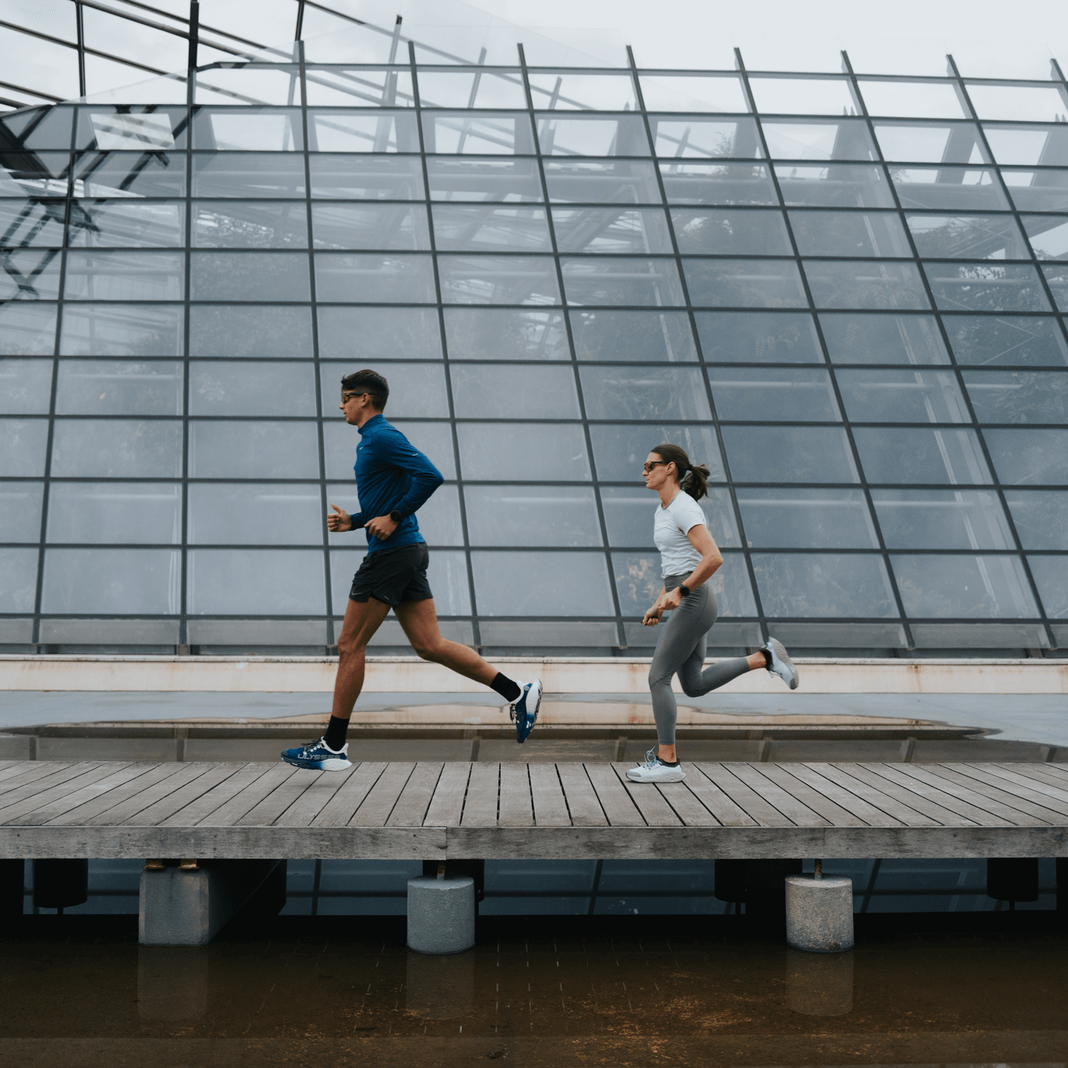 A man and a woman running side by side in a modern architectural environment, showcasing Ulysses Running Shoes from the Road Collection 2025.