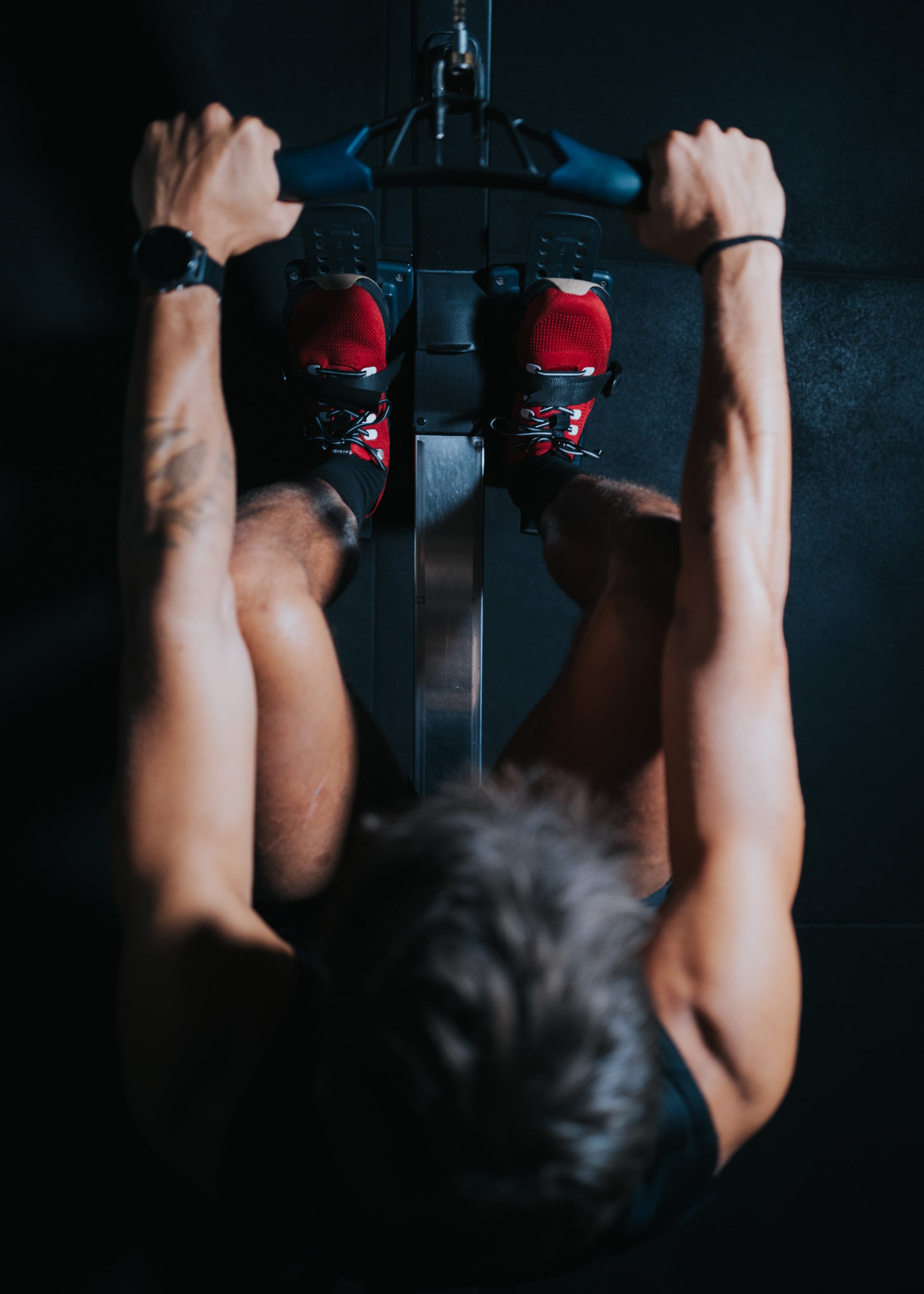 Top-down view of an athlete using a rowing machine, wearing red Ulysses training shoes.