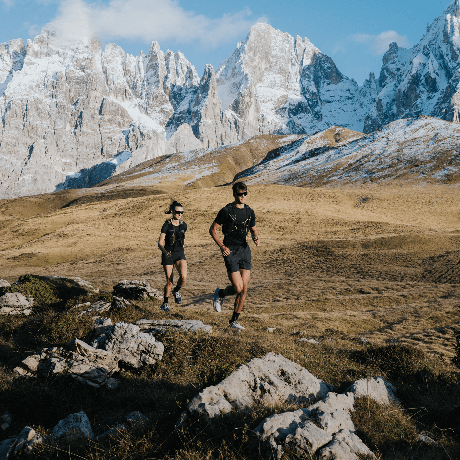 Two trail runners moving across a rocky mountain landscape with snowy peaks in the background, wearing Ulysses Running Shoes from the Trail Collection 2025.