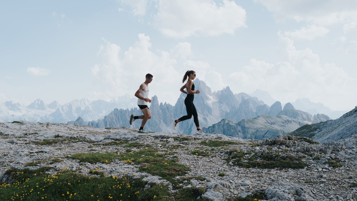 Two runners jogging on a rocky mountain ridge with dramatic peaks and a cloudy sky in the background.