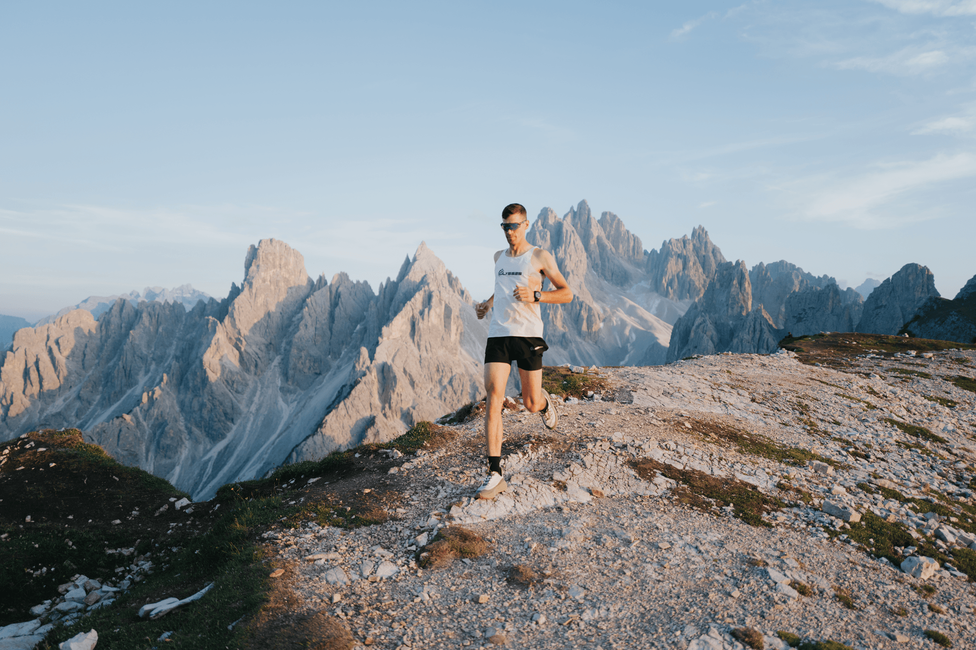 Man trail running along a rocky mountain ridge wearing an Ulysses tank top and shoes, with dramatic jagged Dolomite peaks in the background.