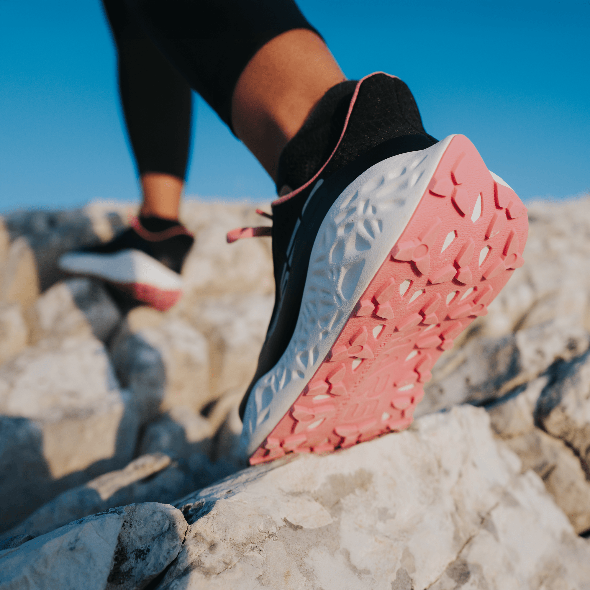 Close-up rear view of the pink and white outsole of an Ulysses running shoe, mid-stride on a rocky mountain trail with a runner in the background.